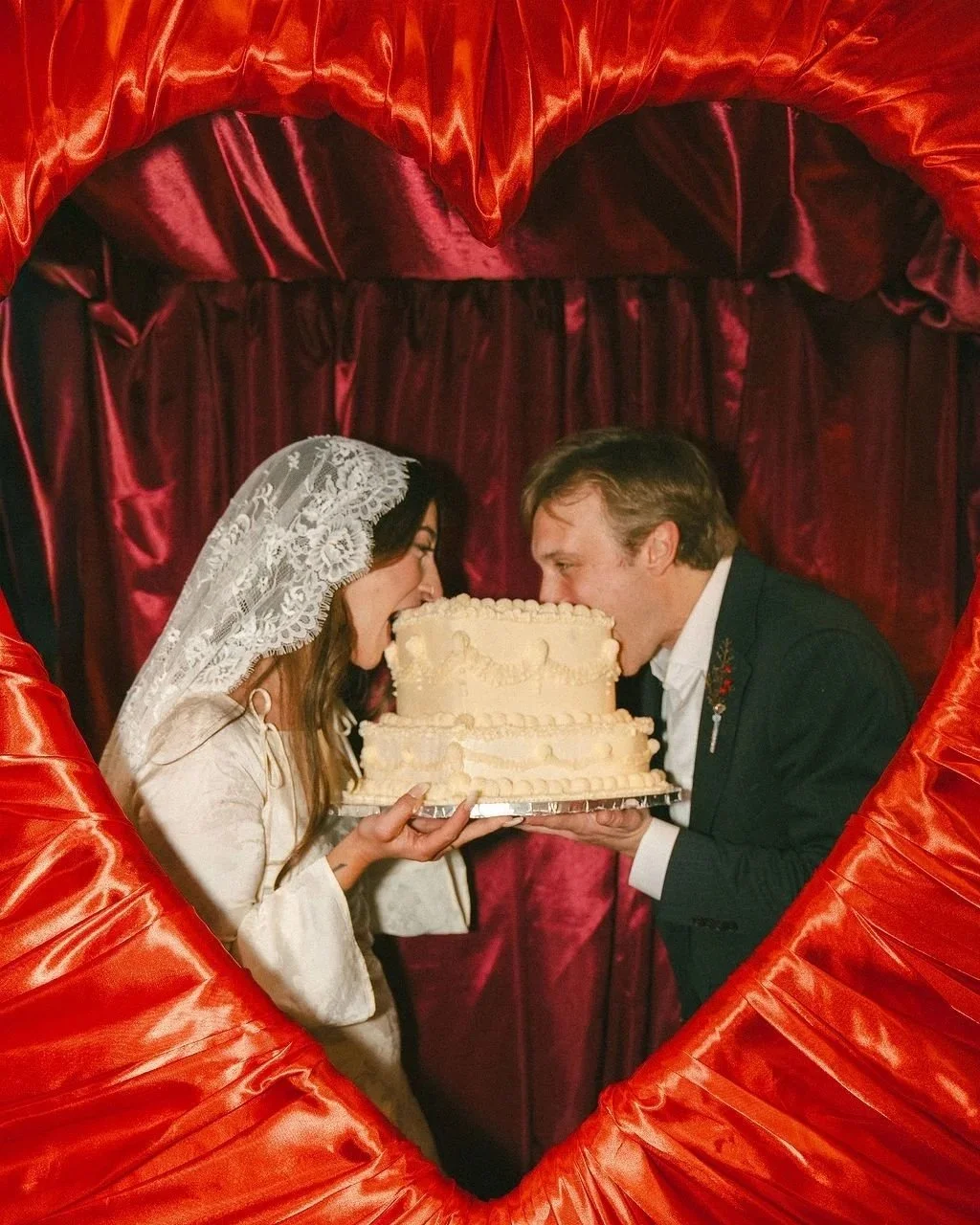 A bride and groom wearing wedding attire biting a large wedding cake together, framed by a red satin heart-shaped backdrop.