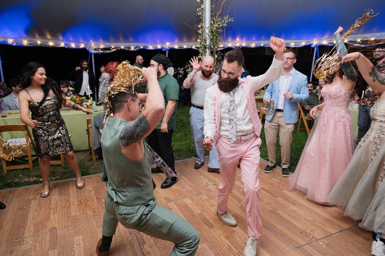People dancing and celebrating at a wedding reception under a canopy decorated with string lights, with some guests throwing golden party streamers.