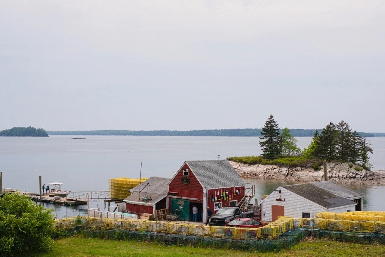 A small waterfront area featuring a red barn-like building, boats, and lobster traps, with a small island in the background and a calm body of water.