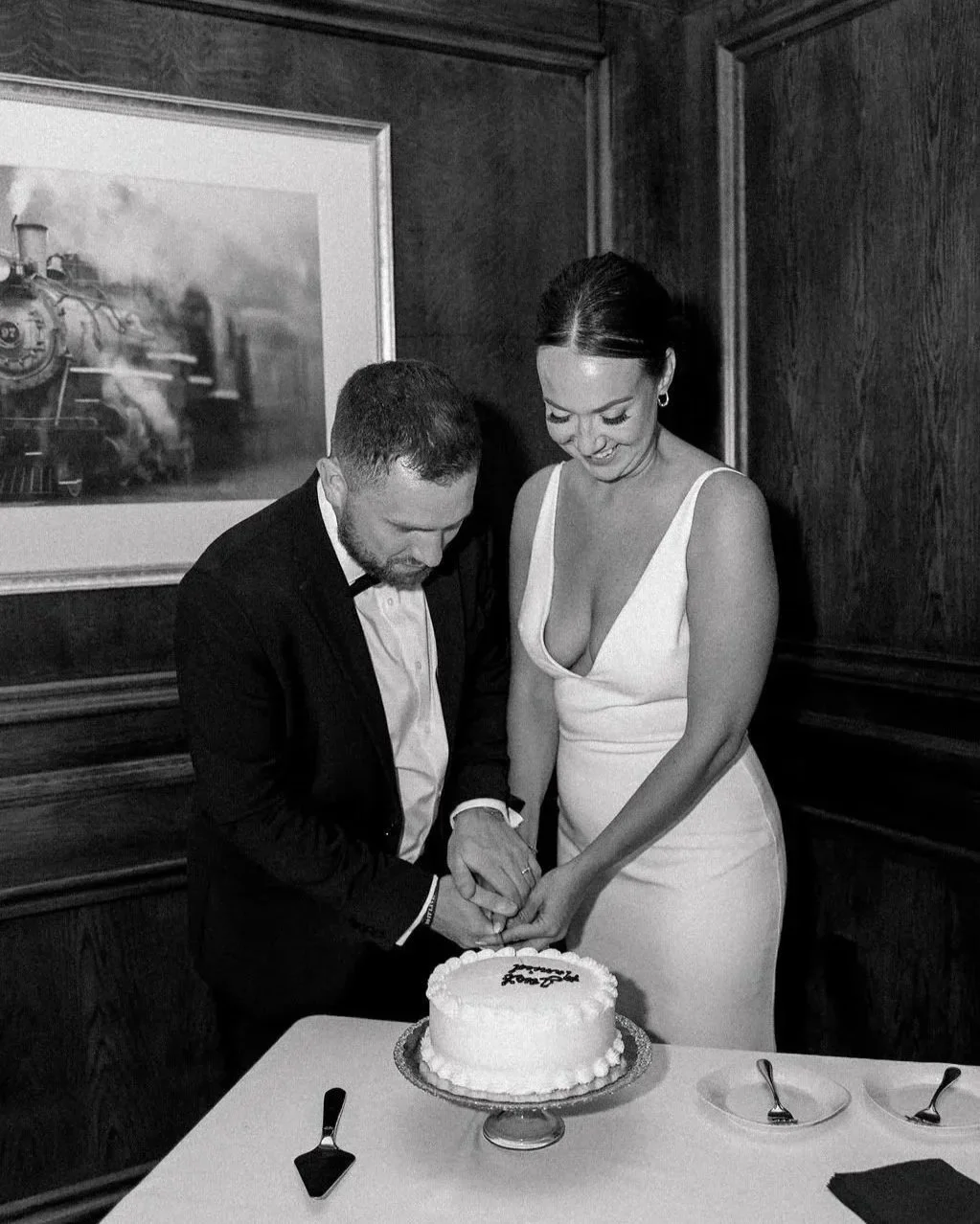 A newlywed couple is cutting a wedding cake at their reception, indoors, in front of a wooden-paneled wall with a framed train picture, in a black and white photo.