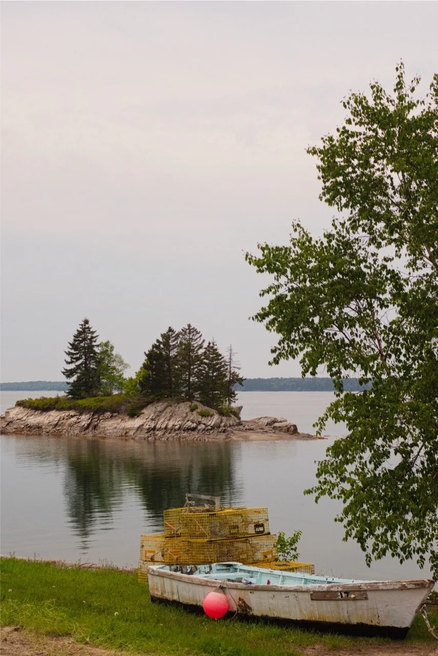 A small boat resting on the grassy shoreline with lobster traps stacked nearby. An island with trees is visible across a calm body of water, under a cloudy sky.