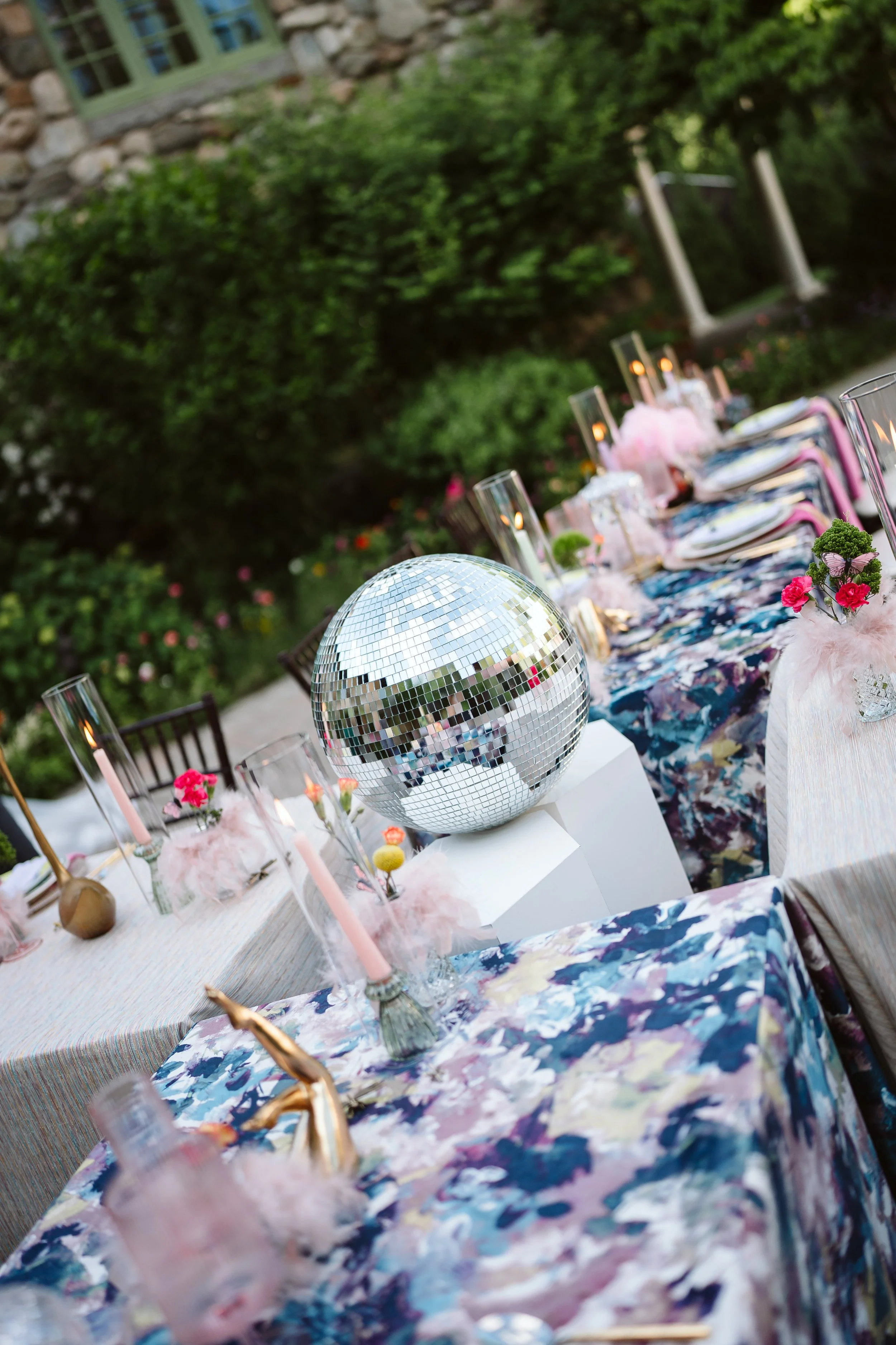 Outdoor dining table decorated for a celebration with a disco ball, pink candles, floral arrangements, and colorful tablecloths, surrounded by greenery and a stone house in the background.