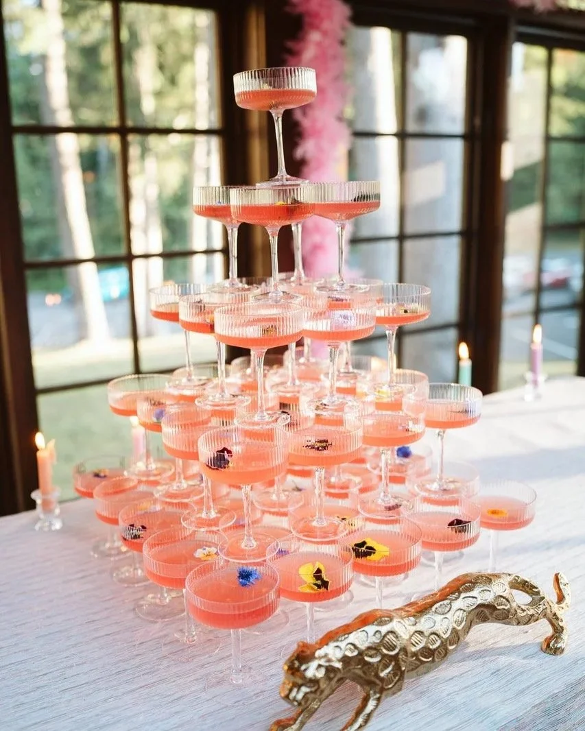 A pyramid of pink cocktails in coupe glasses with edible flowers, set on a white table with candles and a gold panther figurine, in a room with large windows showing trees outside.