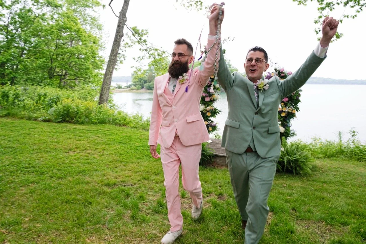 Two men in suits celebrating outdoors by a lake, holding hands in the air with joy, surrounded by green trees and flowers.