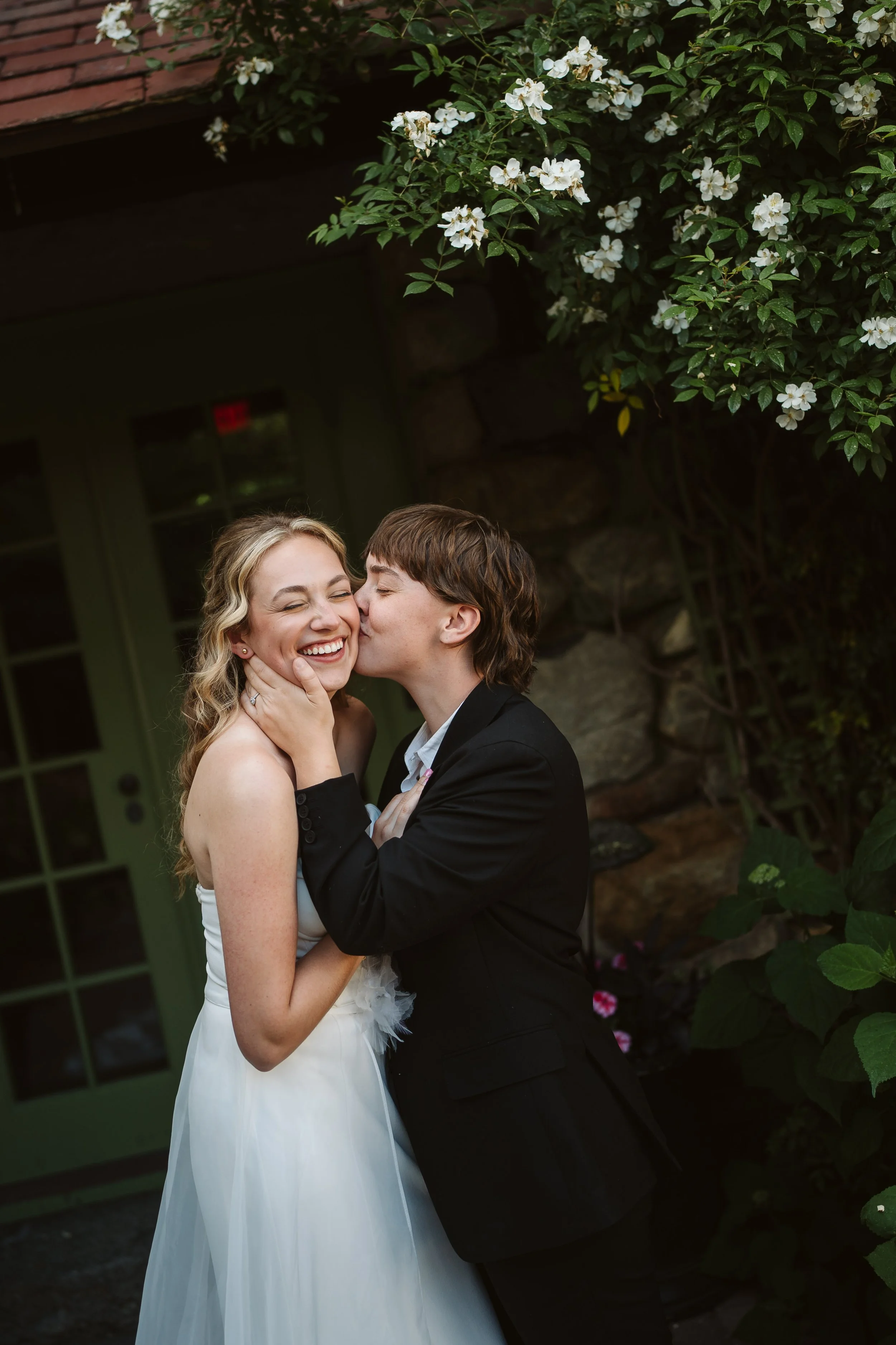 A couple on their wedding day sharing a joyful kiss outdoors, with white flowers and greenery in the background.
