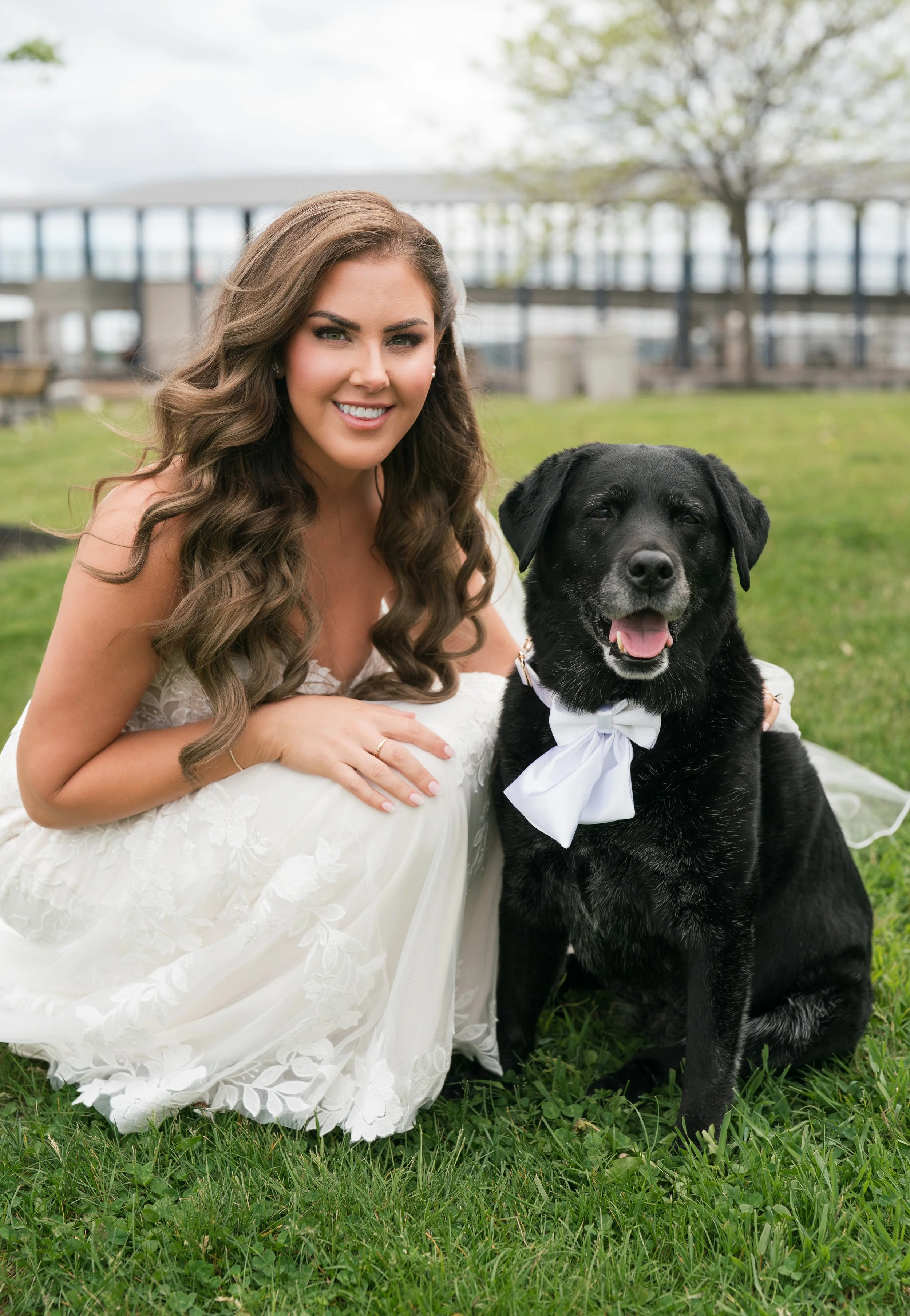 A woman in a white wedding dress with long, wavy brown hair, smiling and laying on grass next to a black retriever dog with a white bow tie, outdoors in a park with trees and a bridge in the background.
