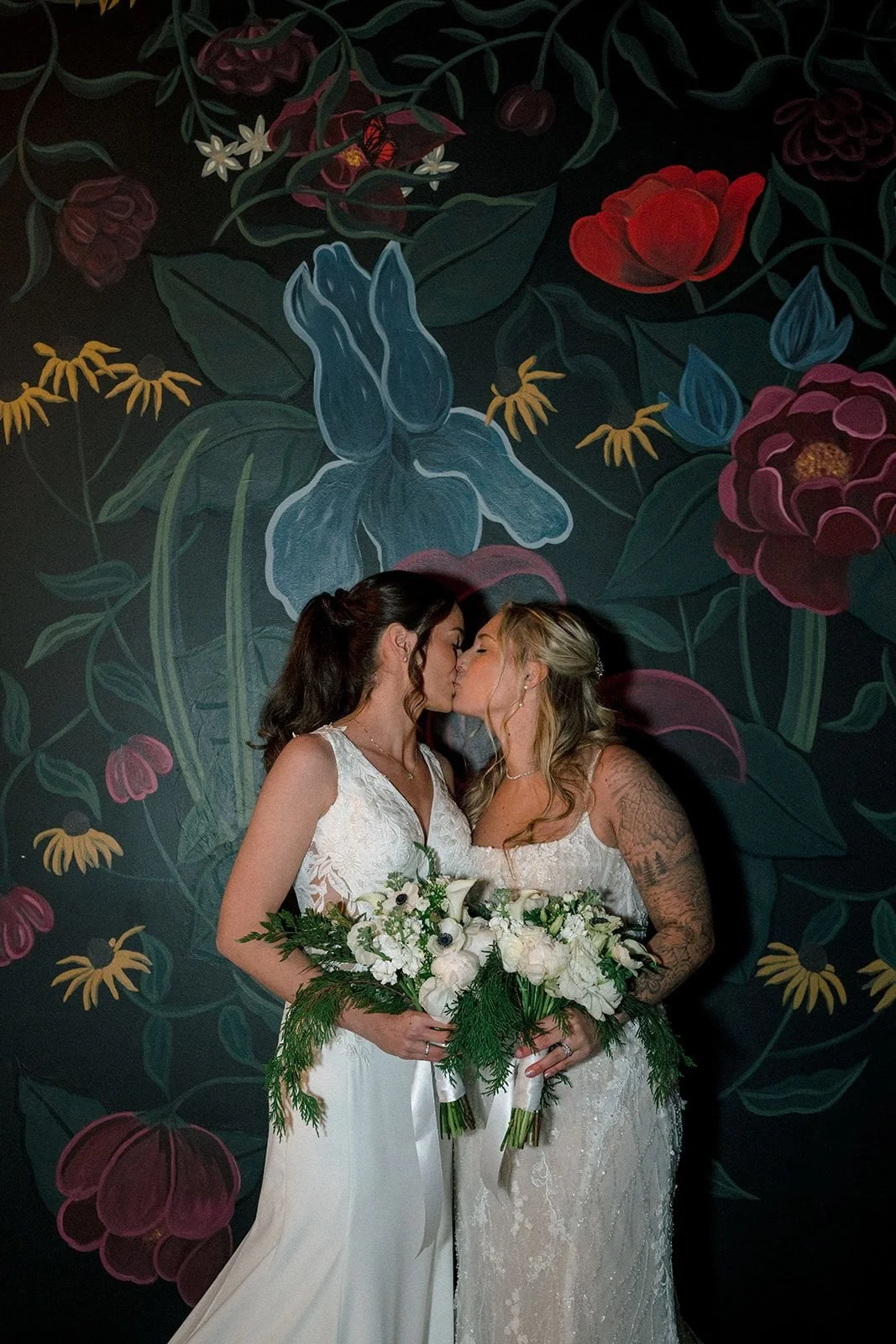 Two women in wedding dresses sharing a kiss while holding bouquets of white flowers, standing against a dark floral mural background.