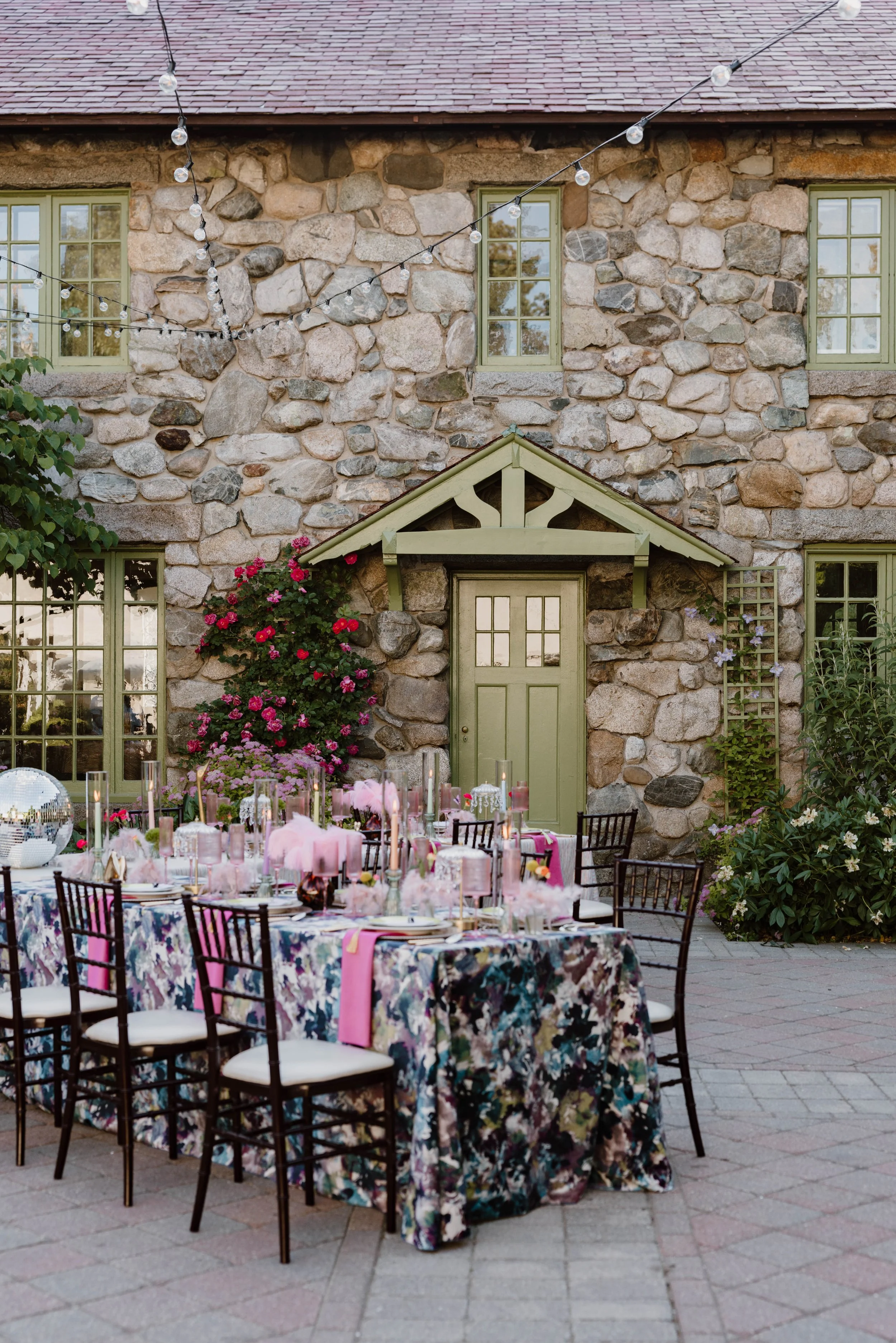 Outdoor dining setup with a long table decorated with a floral tablecloth, pink ribbons, candles, and pink napkins, surrounded by black chairs with white cushions. The background features a stone house with green trim windows and a small porch, decor