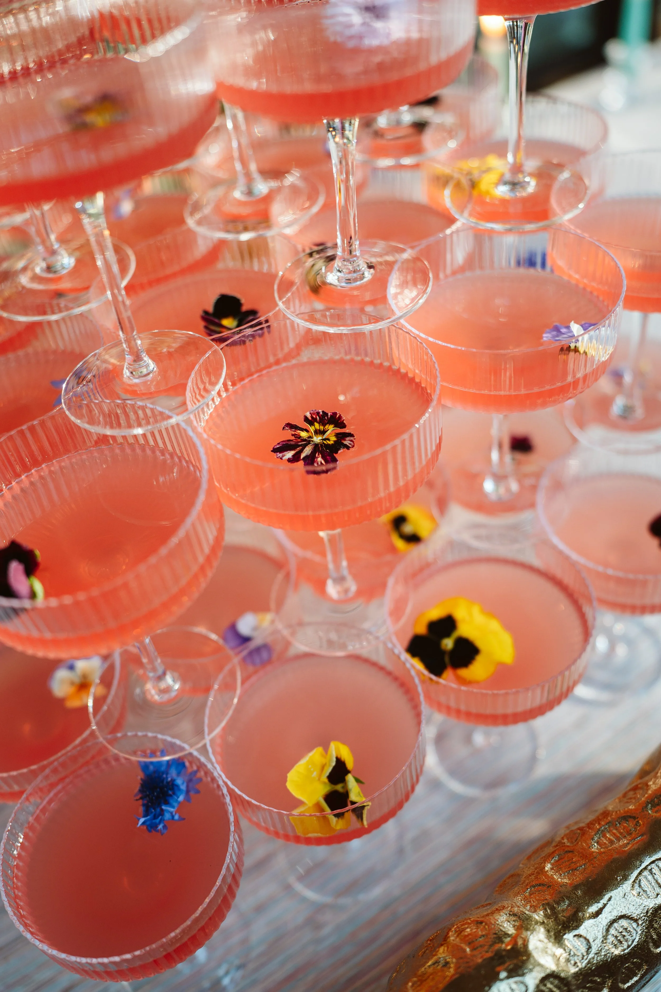 Multiple pink cocktails in martini glasses with edible flowers, arranged in a tiered display at a celebration or event.