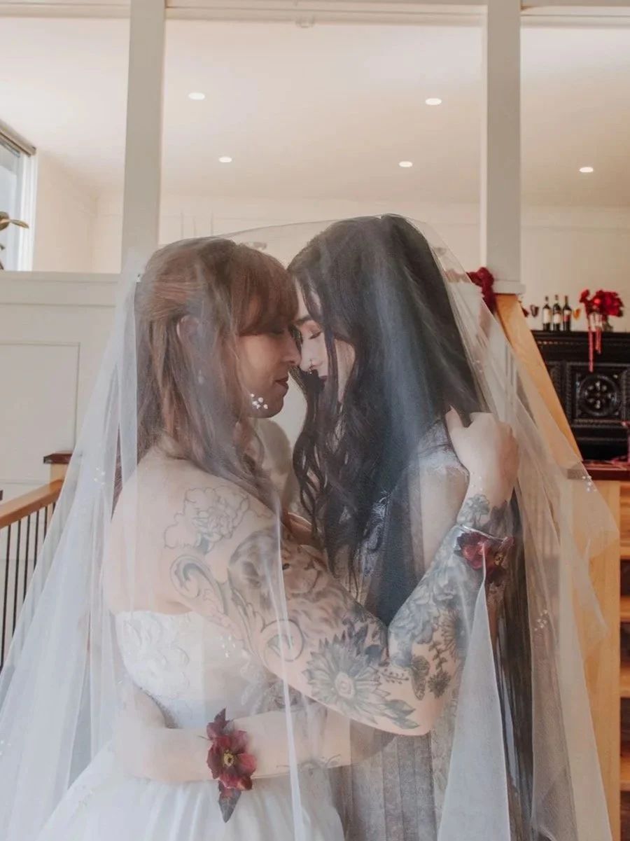 Two women are holding each other close under a veil, touching foreheads, in a wedding setting decorated with flowers and candles.