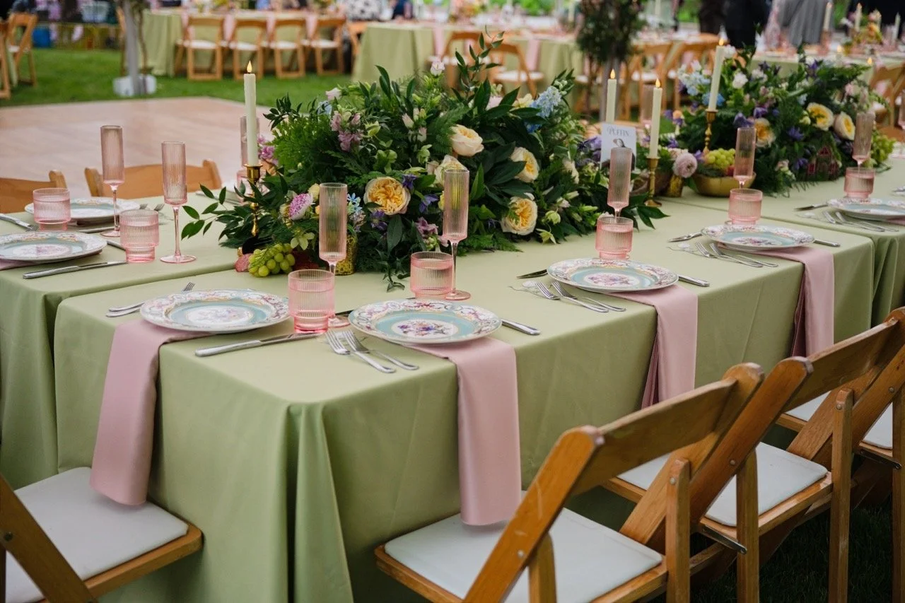 A decorated outdoor dining table with a green tablecloth, pink napkins, floral plates, pink glasses, and a large floral centerpiece, set for a special event.