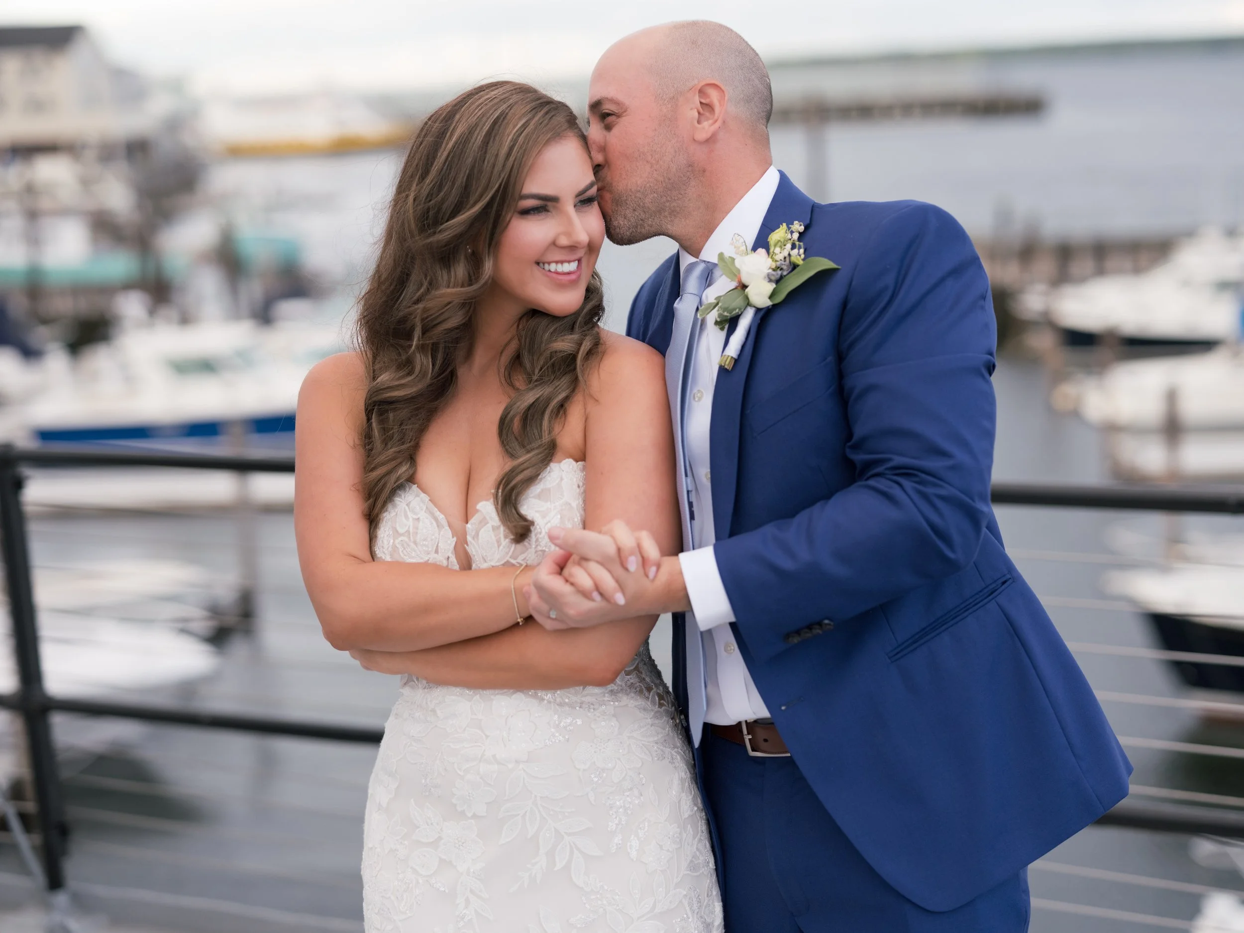 A newlywed couple sharing an intimate moment outdoors near a marina, with boats in the background. The bride is smiling and wearing a white lace wedding dress, and the groom is kissing her on the forehead while dressed in a blue suit with a boutonnie