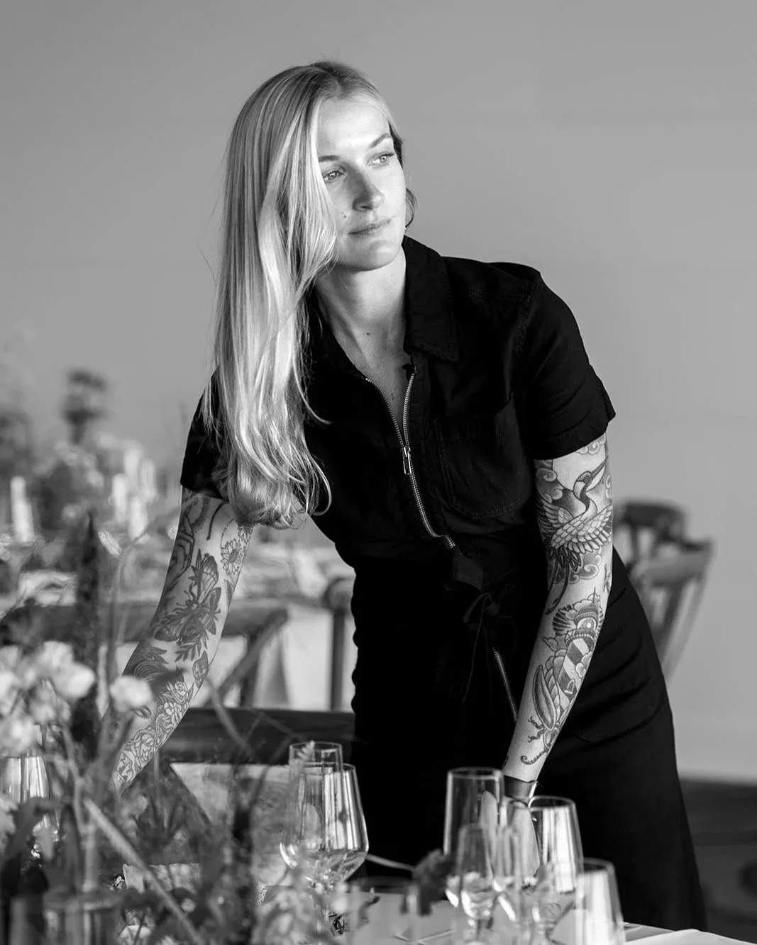 A woman with long blonde hair, tattoos on her arms, wearing a black zip-up outfit, looking thoughtful, standing beside a table with glasses.
