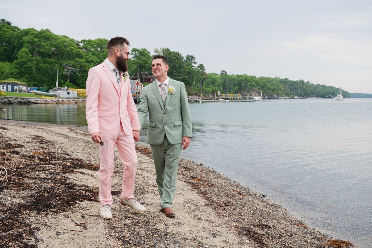 Two men dressed in pastel suits walking on a beach by the water, holding hands, with trees and boats in the background.