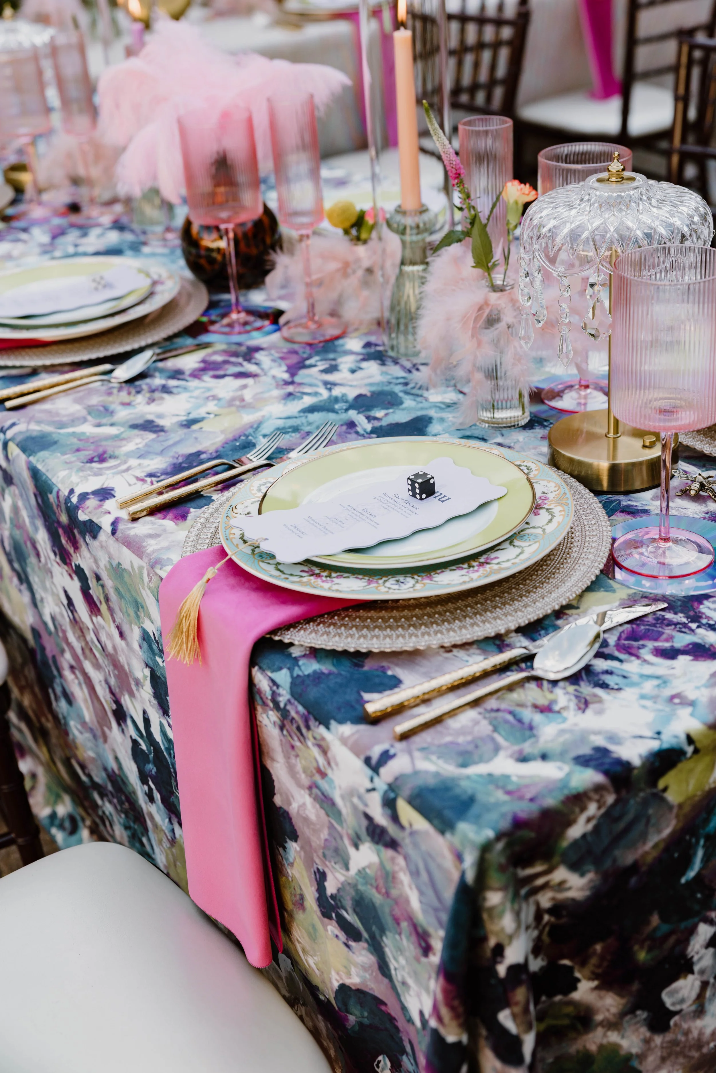 A vividly decorated table featuring a colorful floral tablecloth, pink accents including a pink table runner and pink glassware, a crystal lamp with hanging crystals, and floral centerpieces in glass vases, set for a formal event.