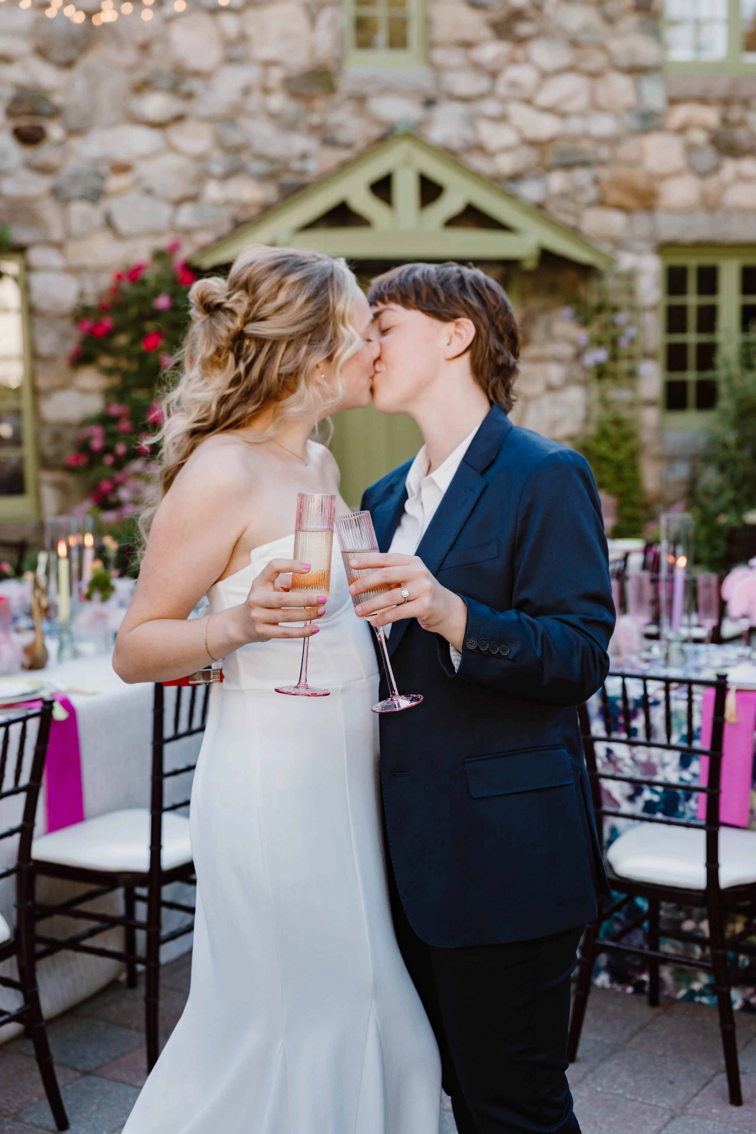 A couple is kissing at a wedding reception outdoors, holding champagne glasses, with decorated tables and chairs in the background.