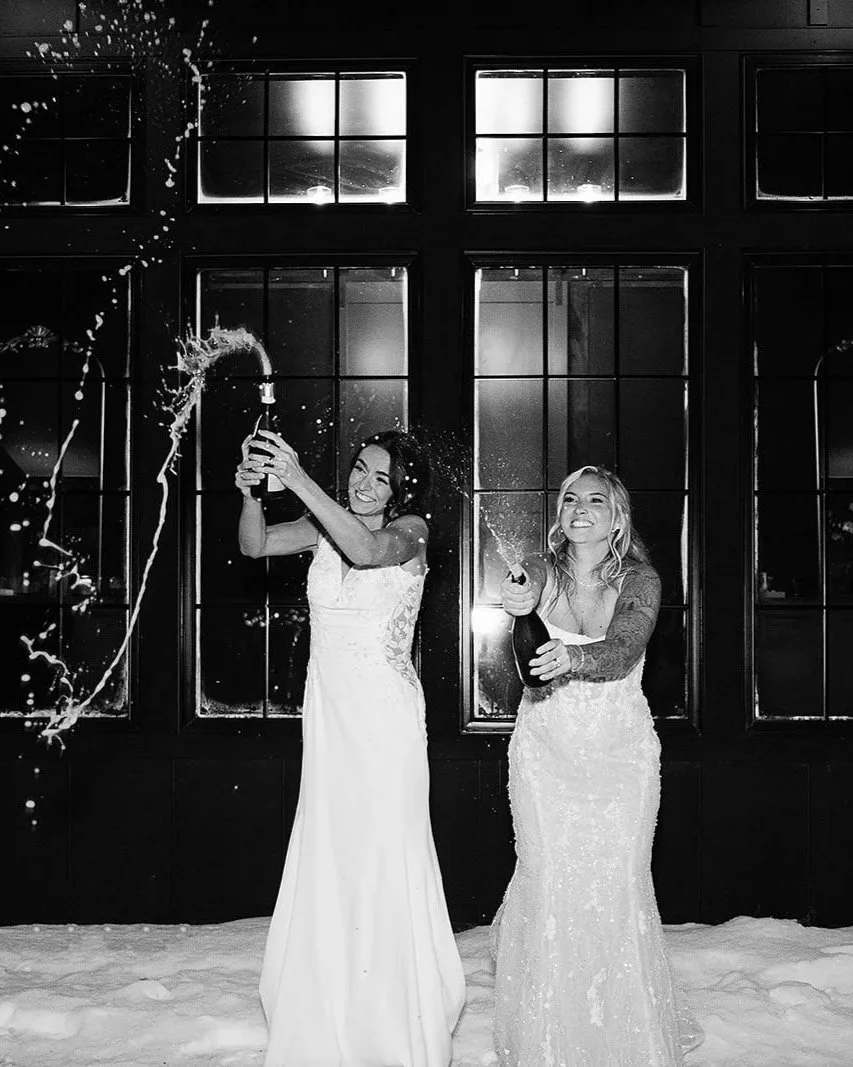 Two women in wedding dresses celebrating with champagne in front of large window at night, with snow on the ground, splashing champagne.