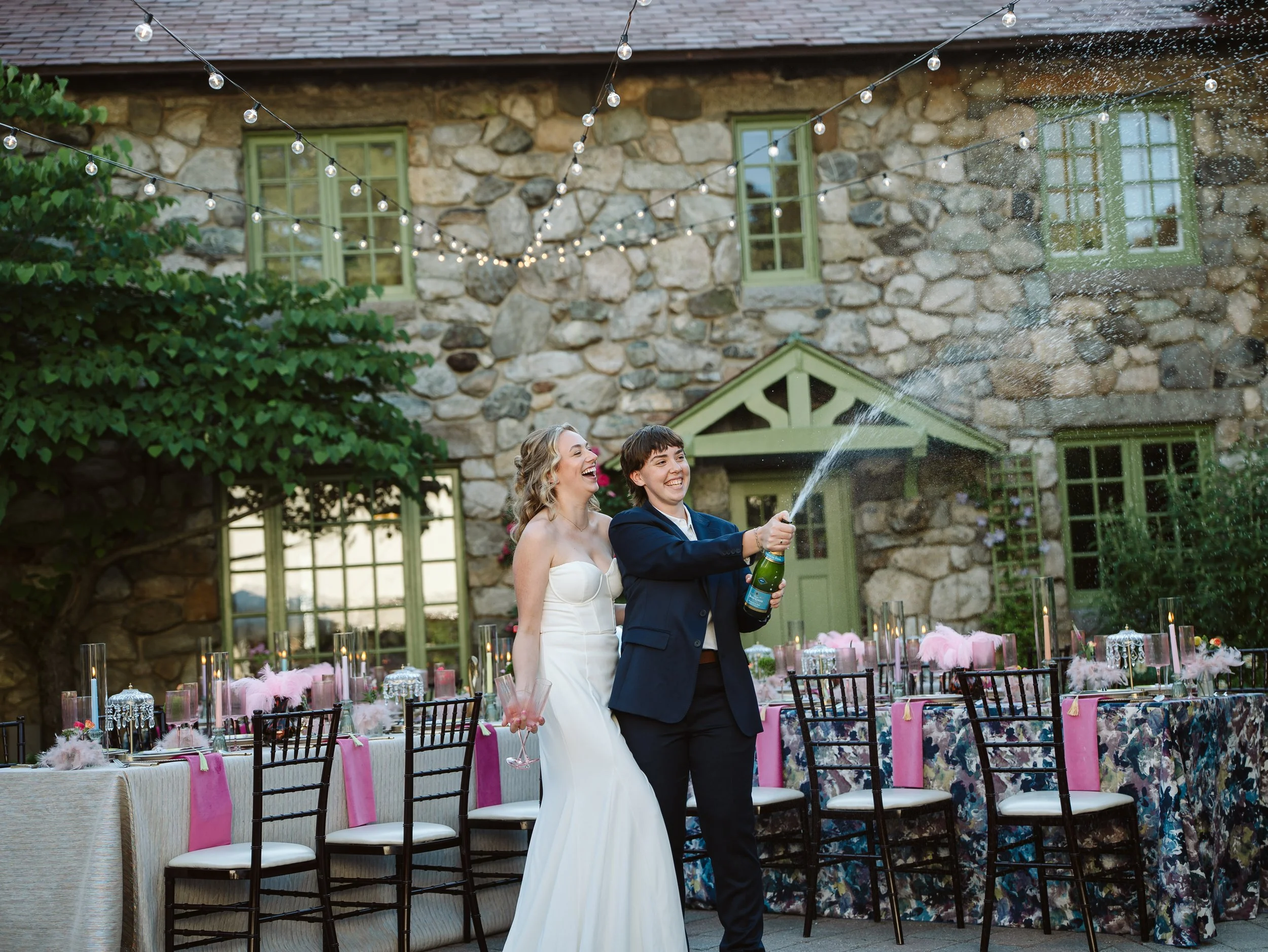 A newlywed couple celebrating at a wedding reception outdoors, with the groom opening a champagne bottle and spraying it, while the bride smiles nearby. The reception area is decorated with pink feathers, candles, and floral arrangements, set against