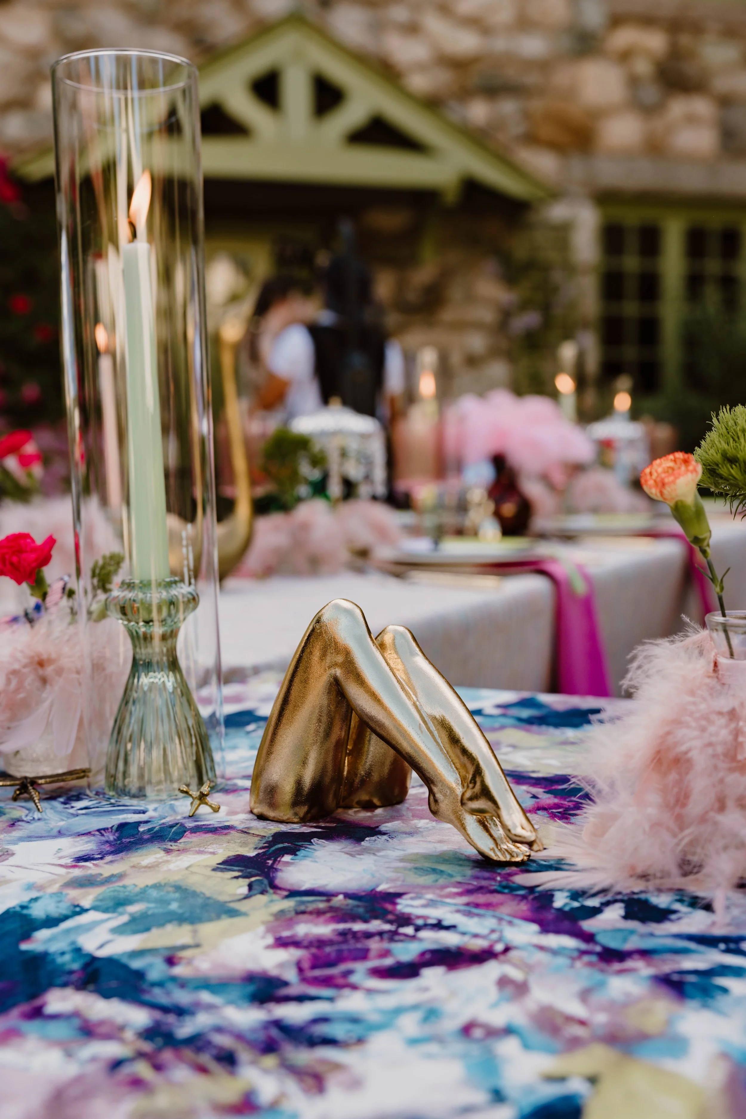 A table centerpiece with a gold high heel shoe, candles, and floral decorations. Out of focus in the background are two people and a house exterior.