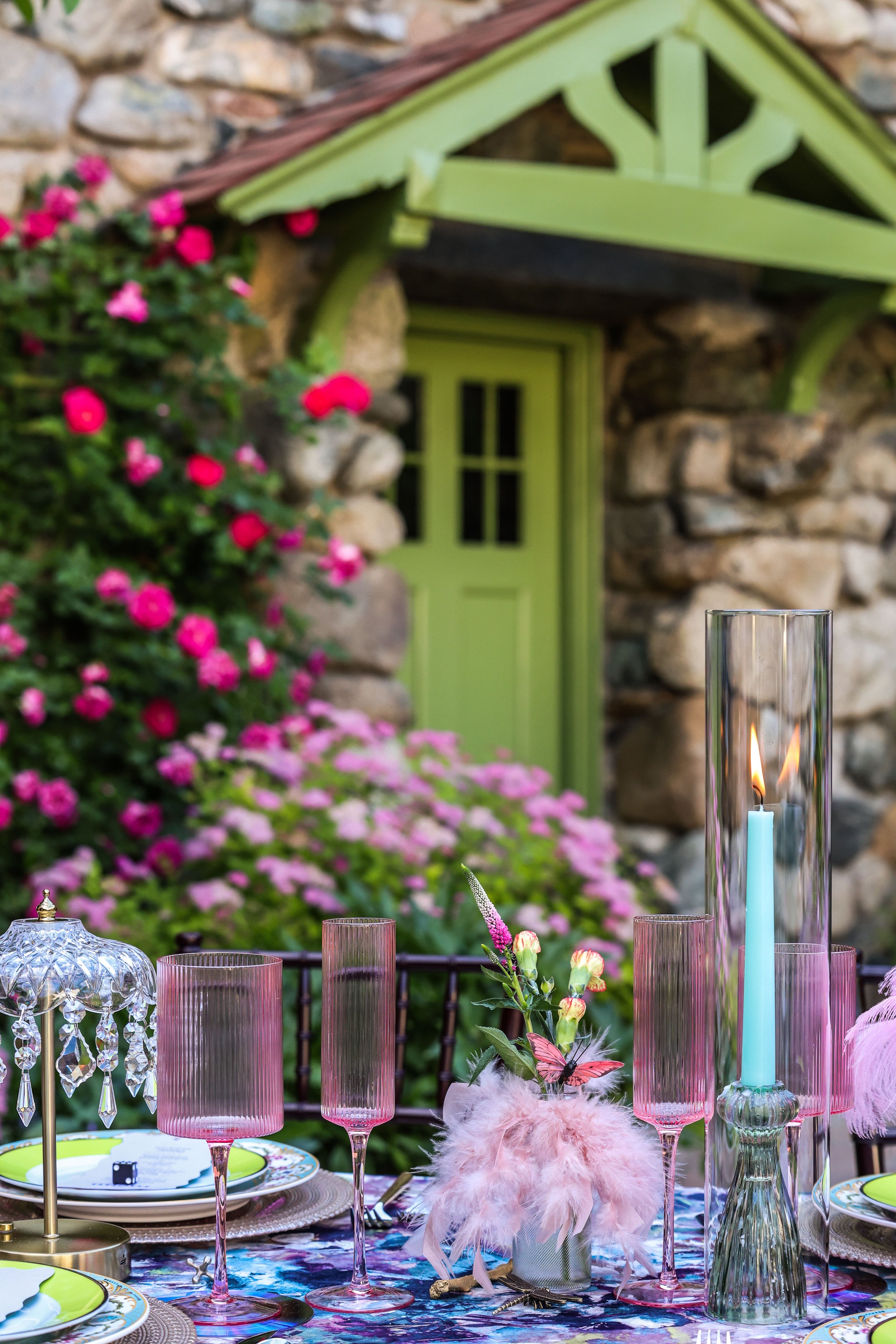 Decorative outdoor table setting with pink glassware, candles, flowers, and a colorful tablecloth, in front of a stone house with a green door and pink flowers.