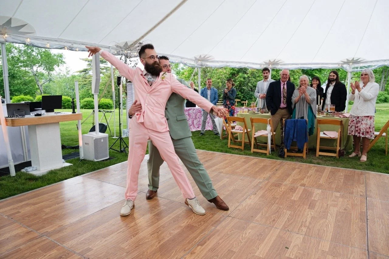 Two men dancing together at an outdoor wedding reception under a white tent, with guests watching and applauding in the background.