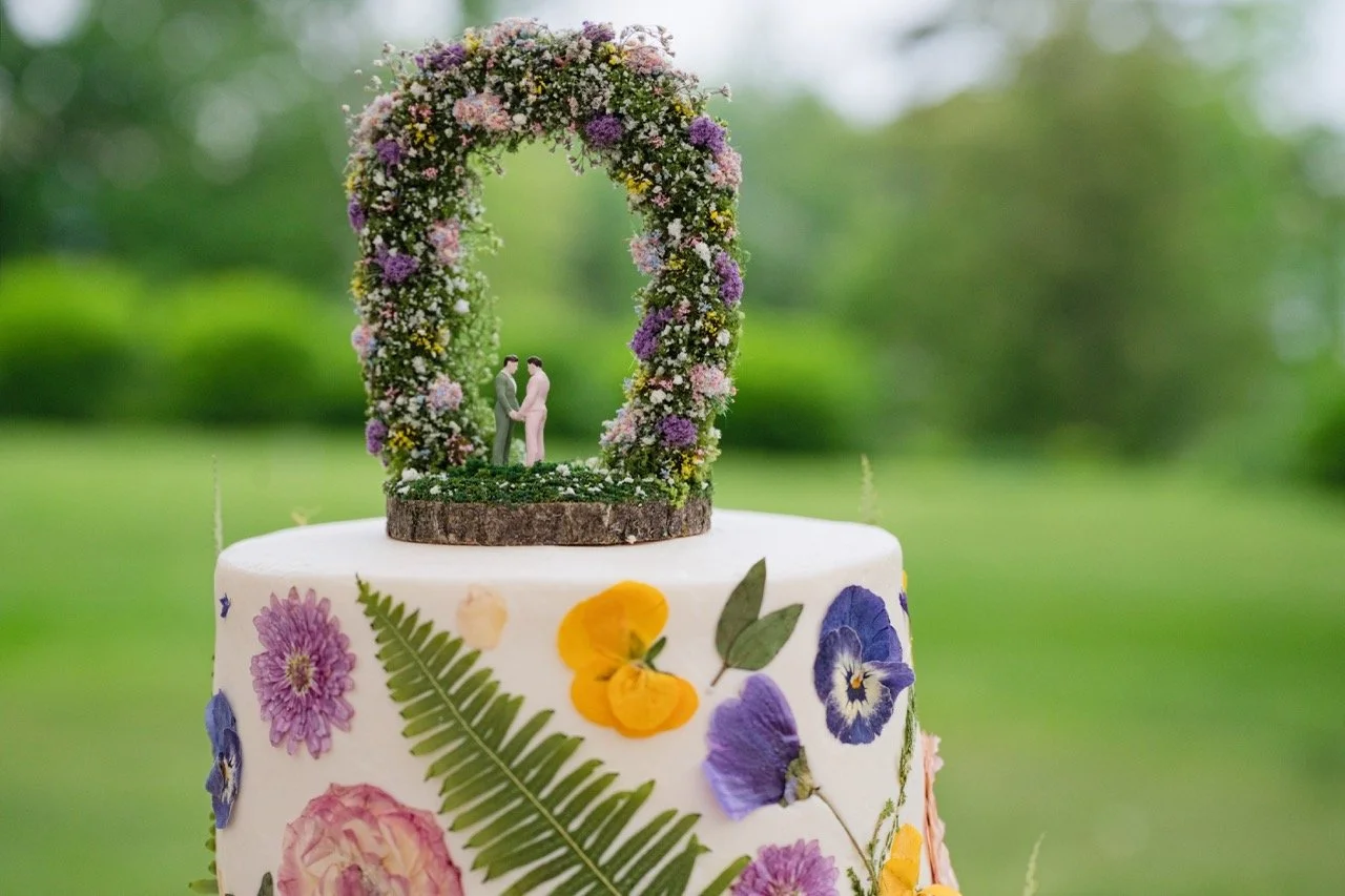 A wedding cake decorated with colorful edible flowers and a floral arch topper with miniature couples holding hands.