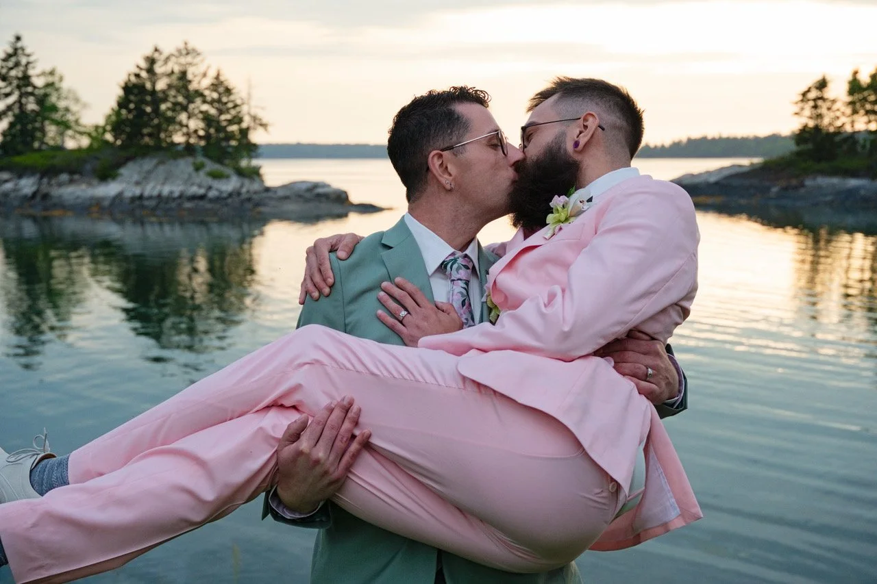 Two men in wedding attire sharing a kiss outdoors near a lake at sunset, one is carrying the other in his arms.