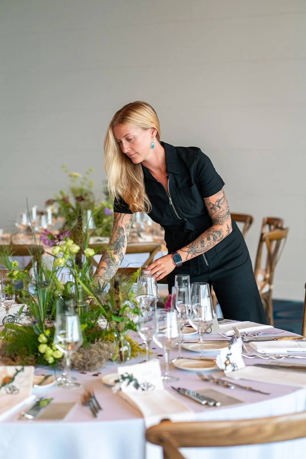 A woman with long blonde hair and tattoos on her arms arranging table settings at a dining event with elegant tableware and floral centerpieces.