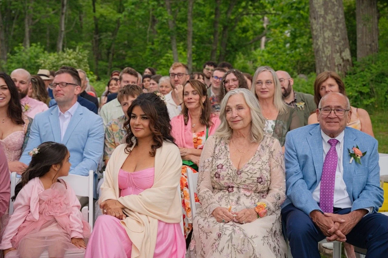 A group of people sitting outdoors in a forested area, attending a wedding ceremony. The crowd includes women and men of various ages, dressed in colorful and formal clothing. They are seated on white chairs and appear to be smiling and enjoying the 