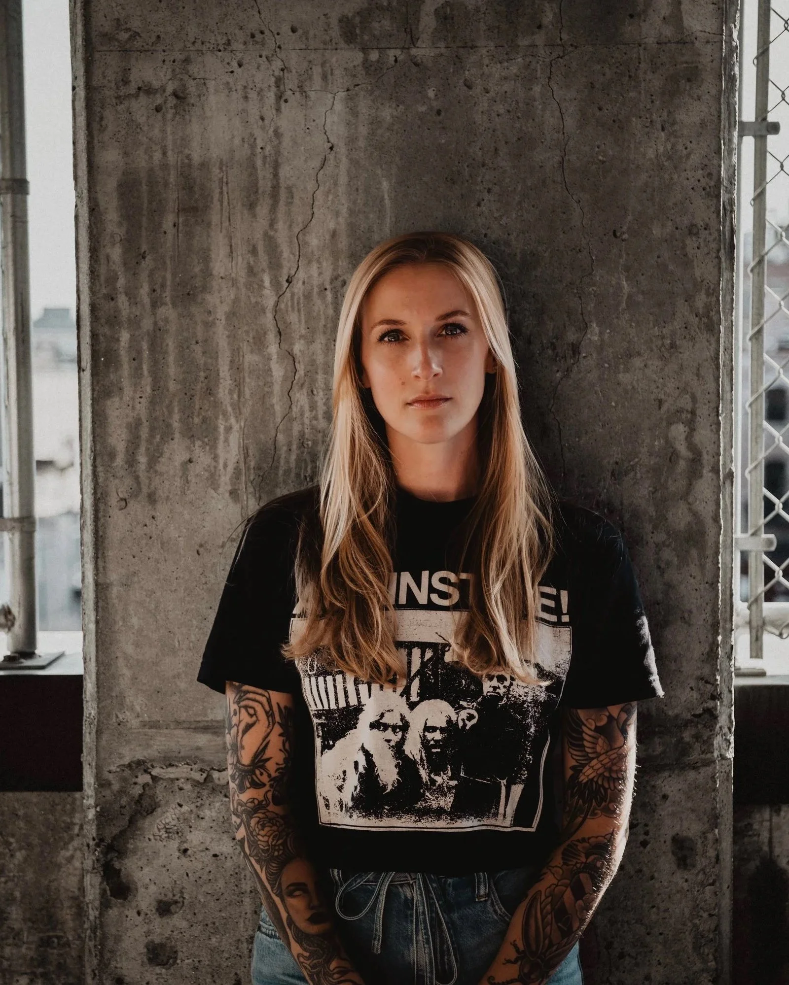 A woman with long blond hair, tattoos on both arms, and wearing a black T-shirt with a graphic print, standing in front of a rough concrete wall with a chain-link fence on either side.
