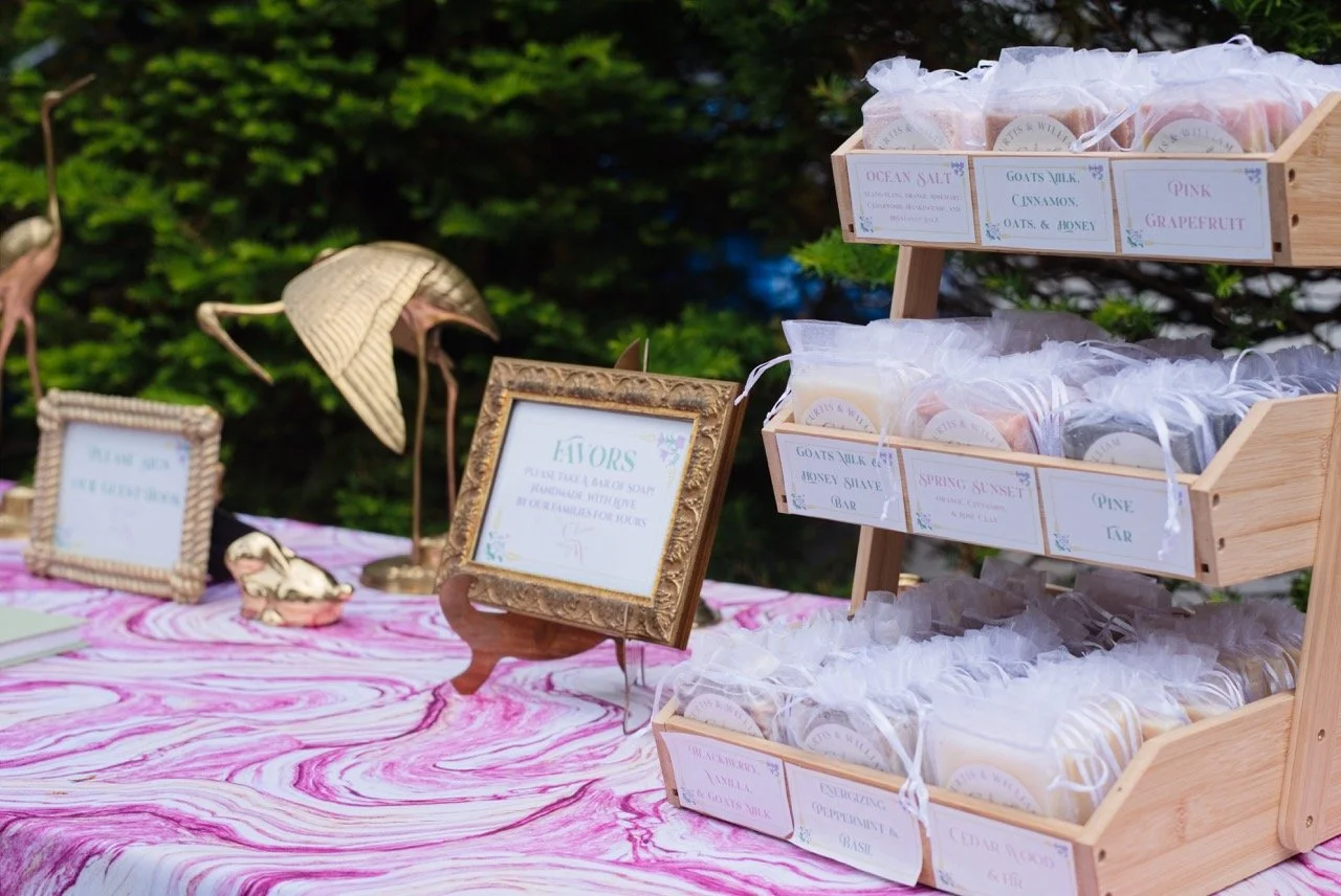 Display of handmade soap bars in a wooden rack at an outdoor market stand, with labels including ocean salt, goat's milk cinnamon, pink grapefruit, honey shave, spring sunset, pine tar, energizing cucumber basil, and cedar woods.