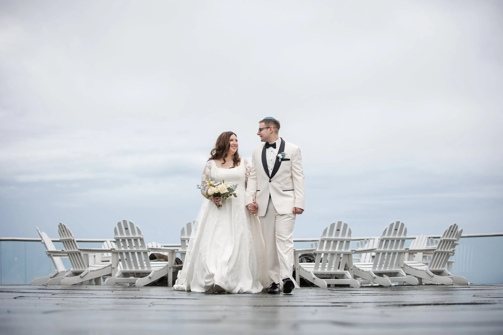 A bride and groom standing hand in hand outdoors on a wooden platform, with white chairs lined up behind them, under an overcast sky, during their wedding celebration.