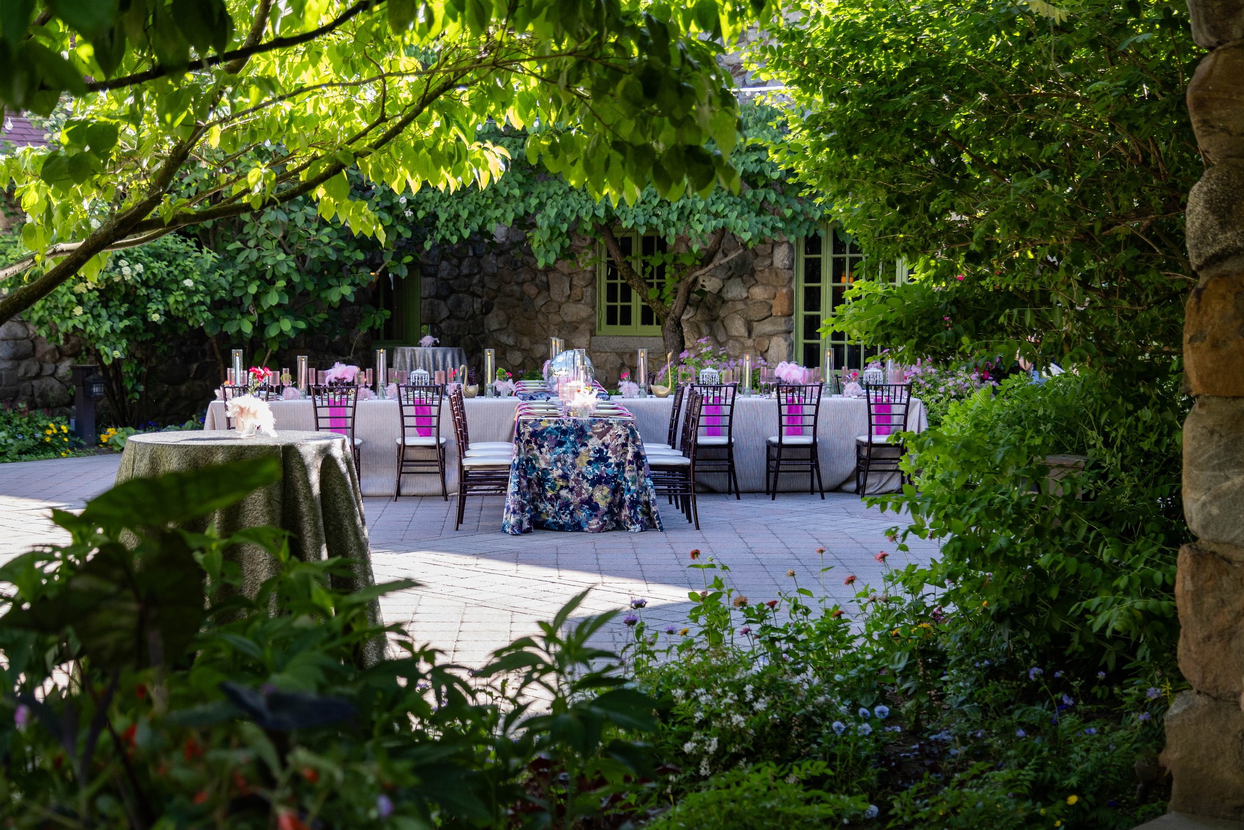 Outdoor garden with a long dining table decorated with pink flowers and candles, surrounded by chairs, and a stone house covered in green ivy in the background, shading the scene with trees.