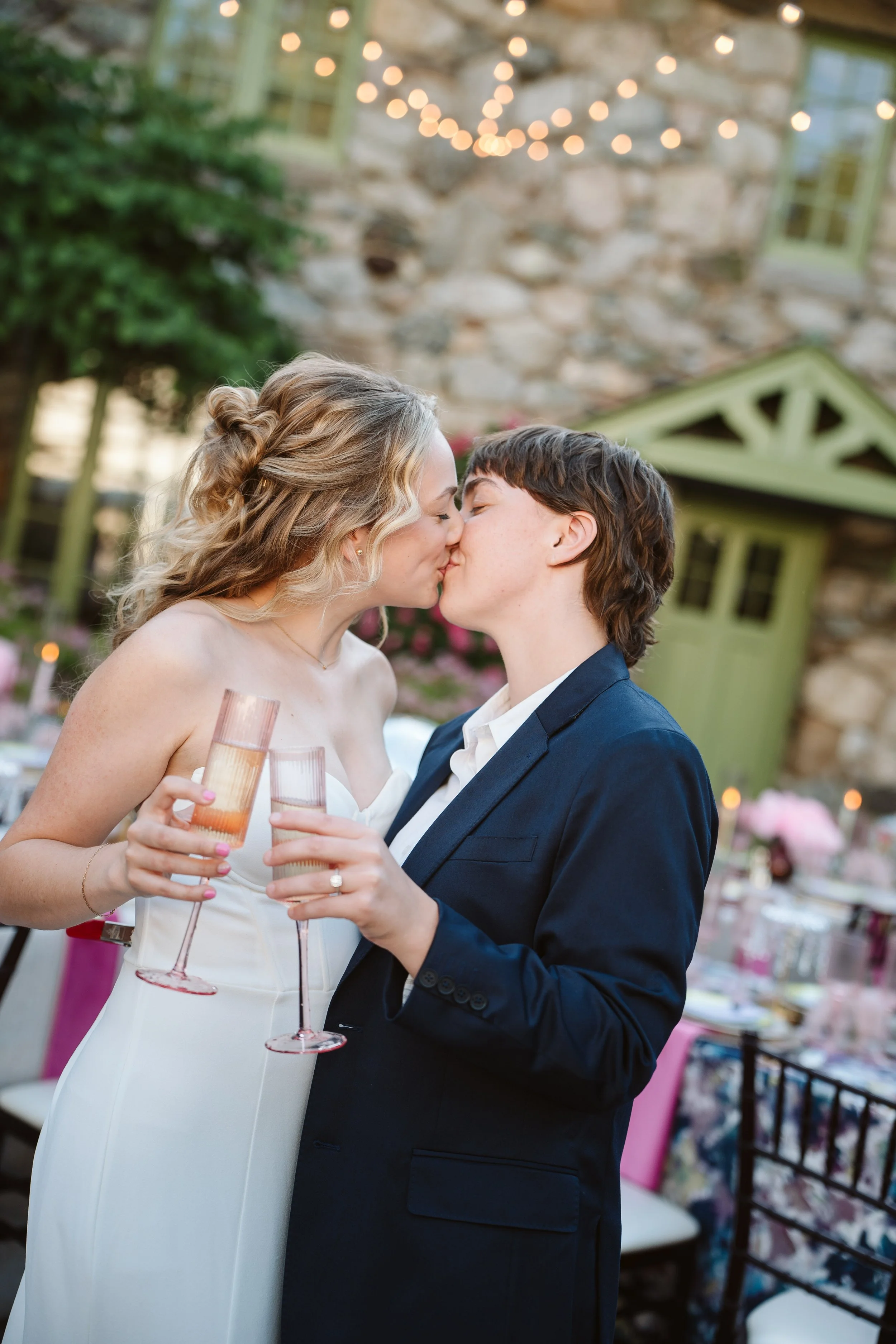 A couple sharing a kiss at a wedding reception, holding pink champagne glasses, with a stone wall and string lights in the background.
