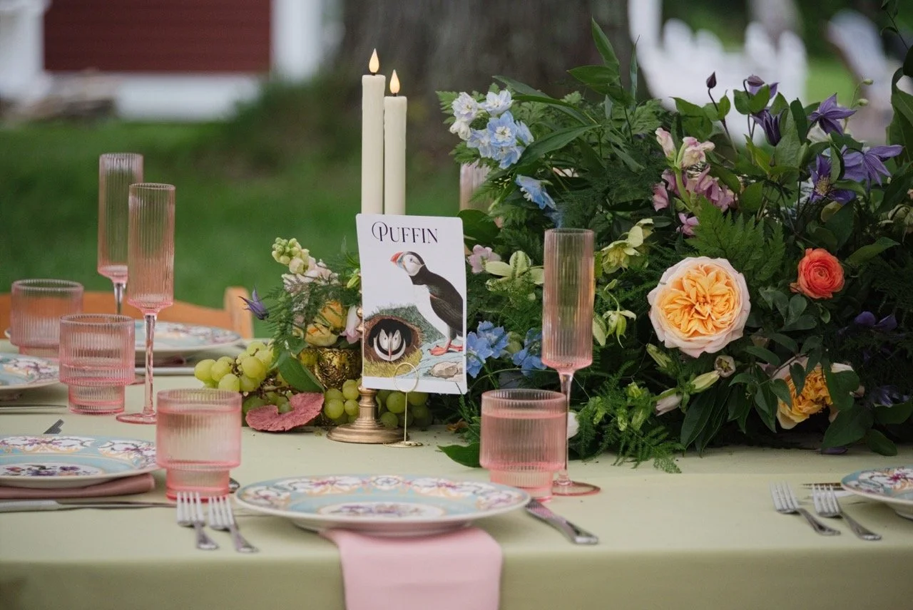 Table decorated with pink glasses, floral plates, and a floral centerpiece featuring various flowers and greenery. A menu card with a puffin illustration is also part of the table setting.