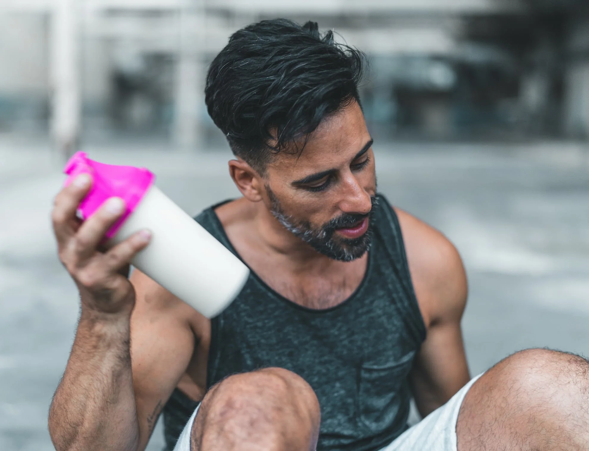 Man sitting outdoors in athletic clothing, holding a white water bottle with a pink cap, with a serious or tired expression.