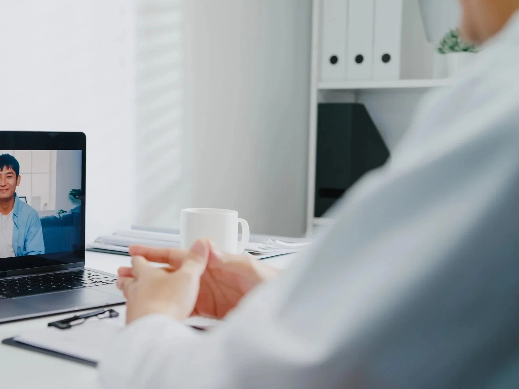 Person participating in a video call on a laptop, sitting at a desk with a white coffee mug, in a bright, organized office setting.
