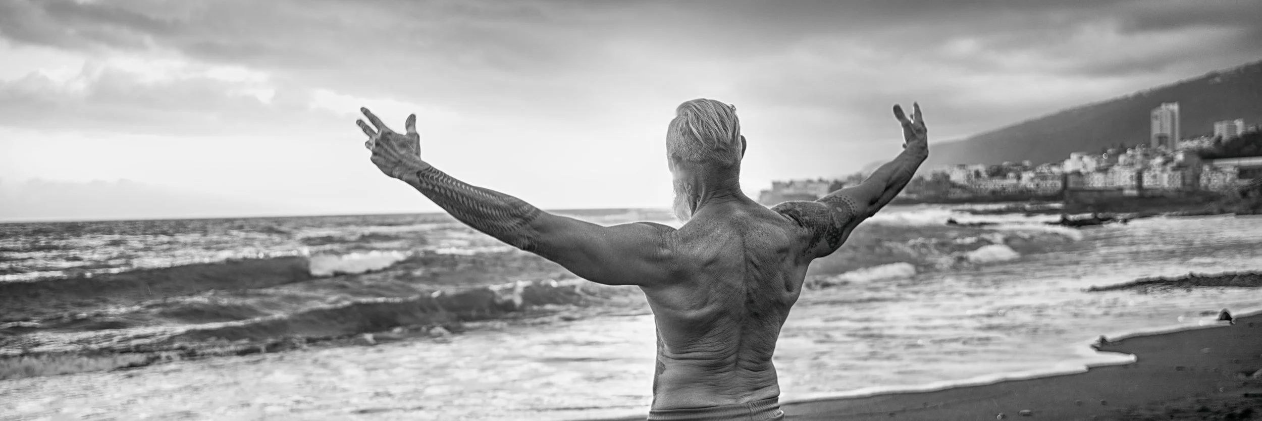 A tattooed shirtless man with gray hair standing on a beach with his arms outstretched, facing the ocean and a cloudy sky, with a cityscape and mountains in the distance.
