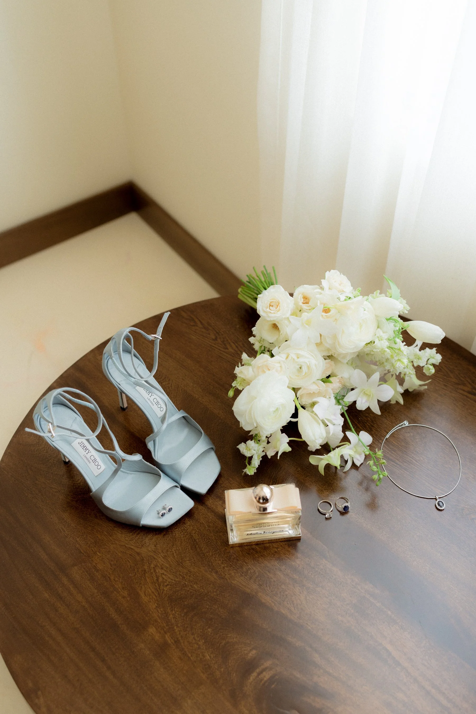Zapatos de tacón en color gris claro, reloj, anillos, perfume y un ramo de flores blancas sobre una mesa de madera, en un entorno iluminado por luz natural.