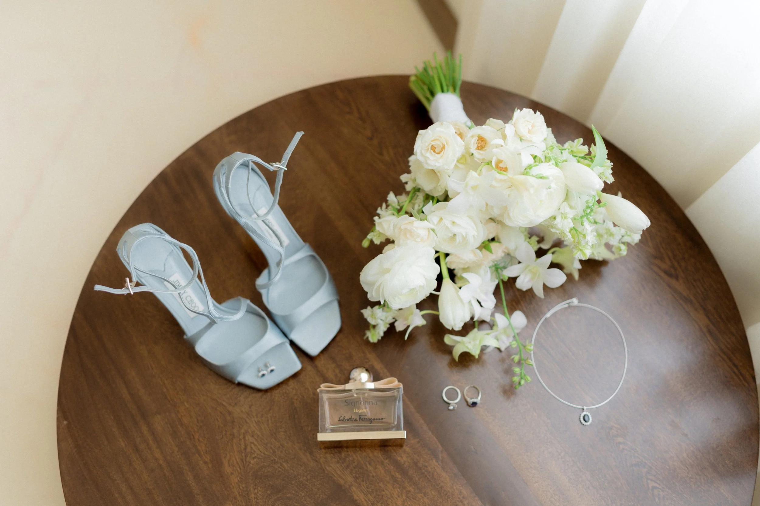 Zapatos de novia azul, ramo de flores blancas, anillos, collar y pendiente en una mesa de madera.