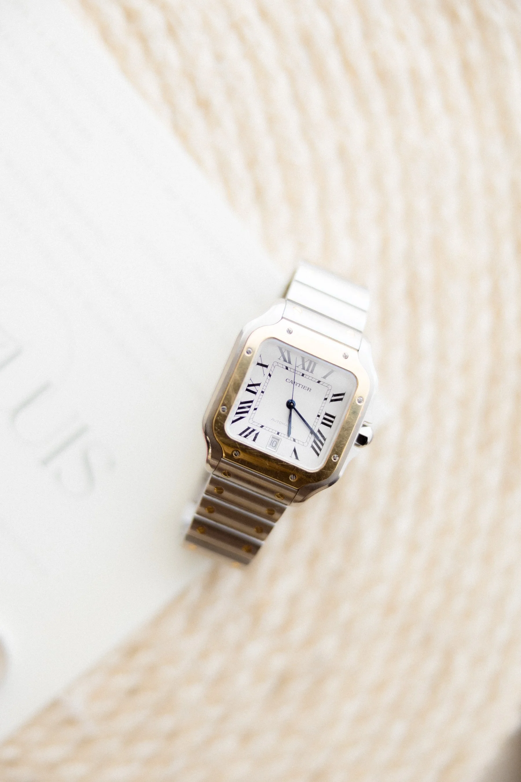 Reloj de pulsera de marca Cartier con caja de acero y detalles en oro, mostrando la hora con marcadores de números romanos, colocado sobre una superficie de madera y un libro.