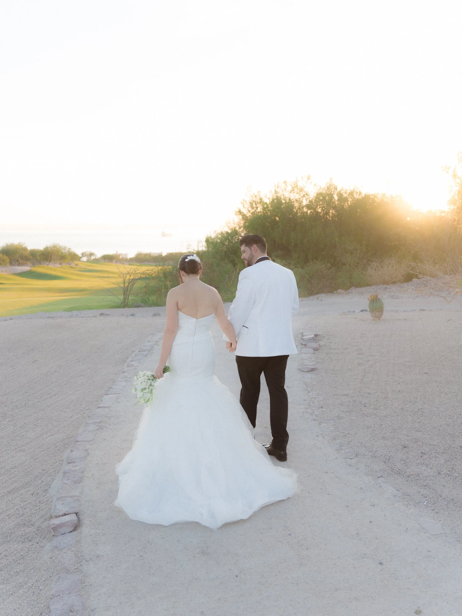 Pareja de novios caminando juntos al atardecer en un paisaje al aire libre, con la novia en vestido de boda y el novio en traje blanco y negro.