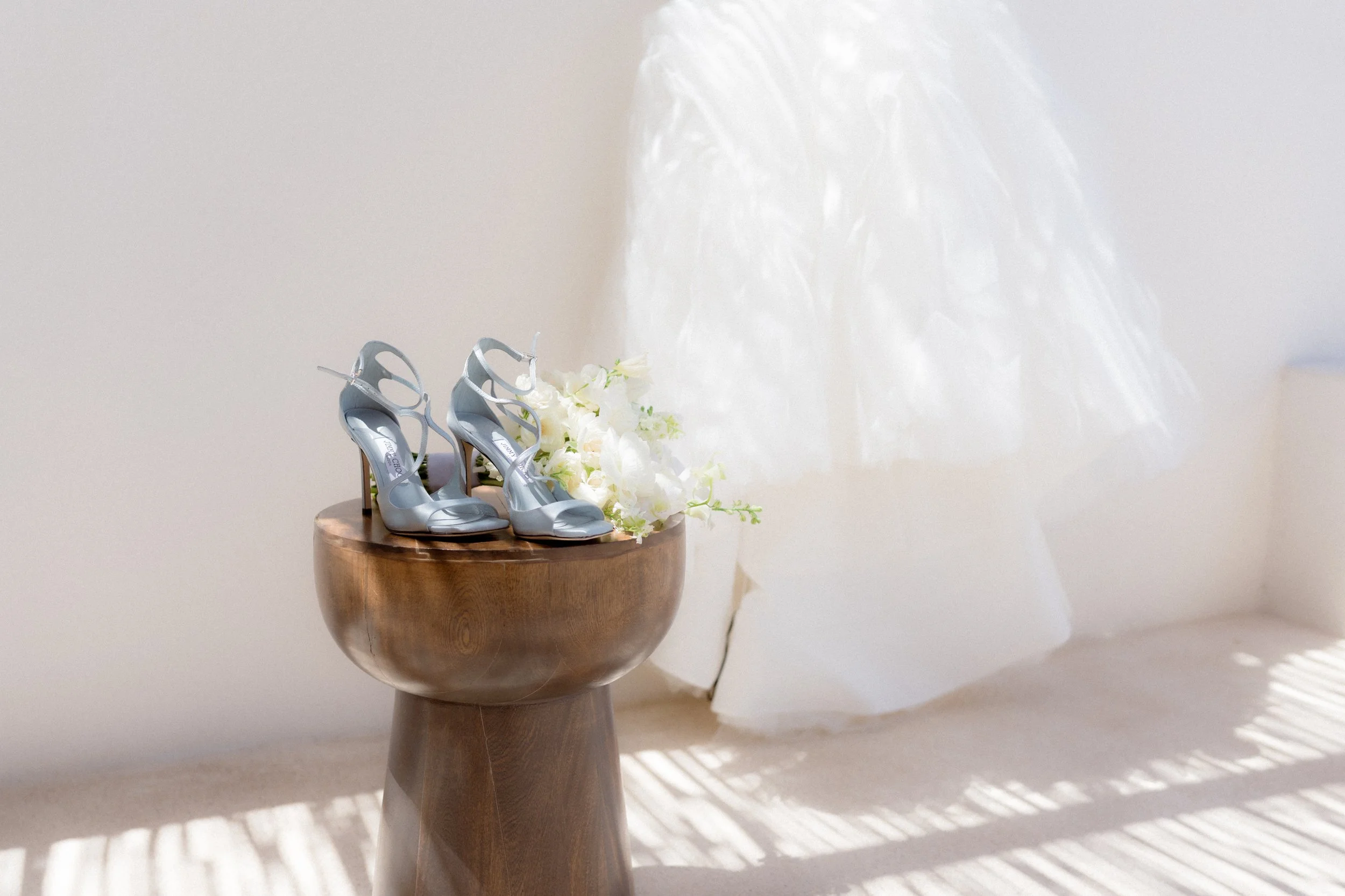 Zapatos de boda plateados junto a un ramo de flores blancas sobre un taburete de madera en un ambiente rústico y brillante.