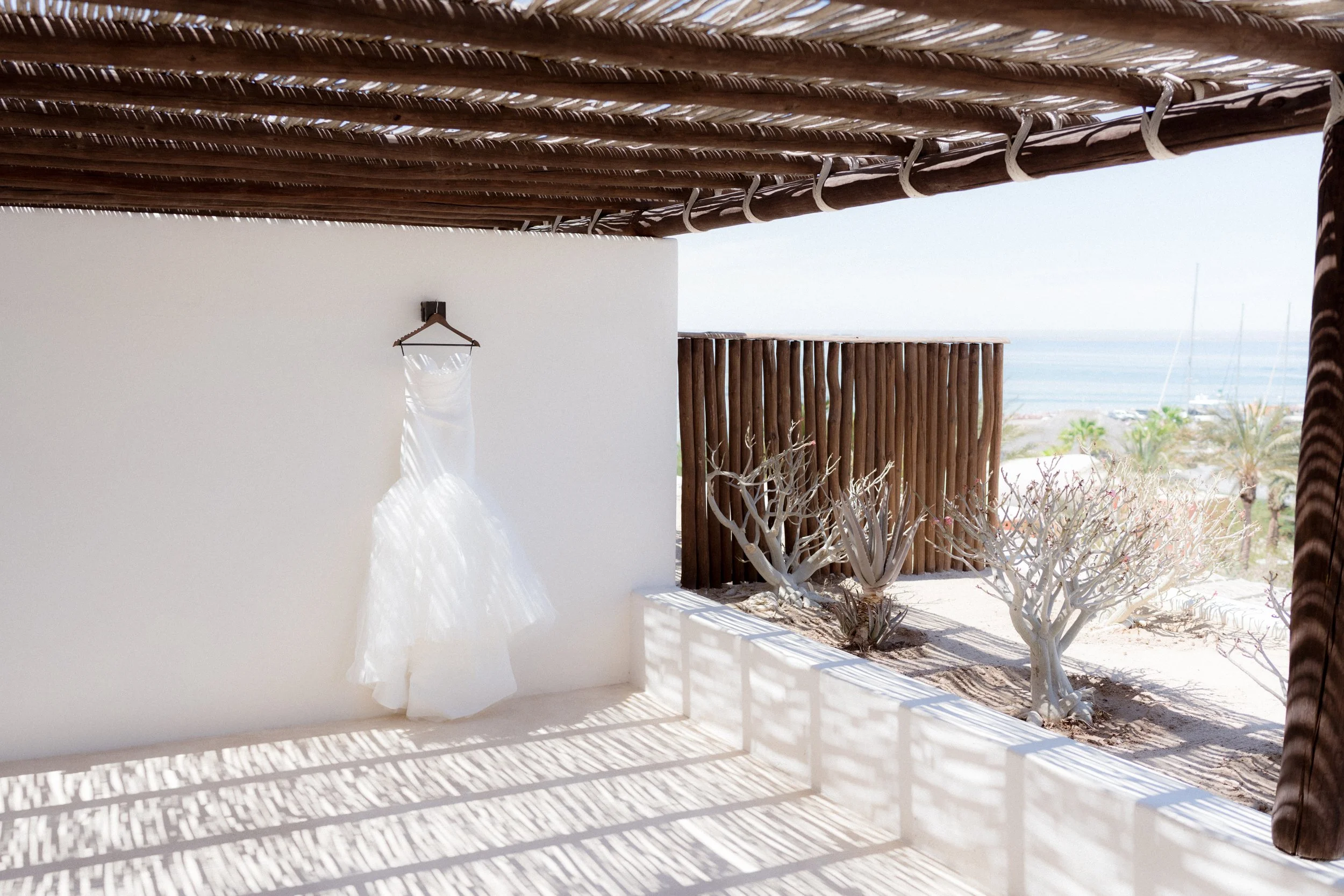 Vestido de novia colgado en una pared blanca en una terraza con sombra y vista al mar y jardines áridos con cactus.
