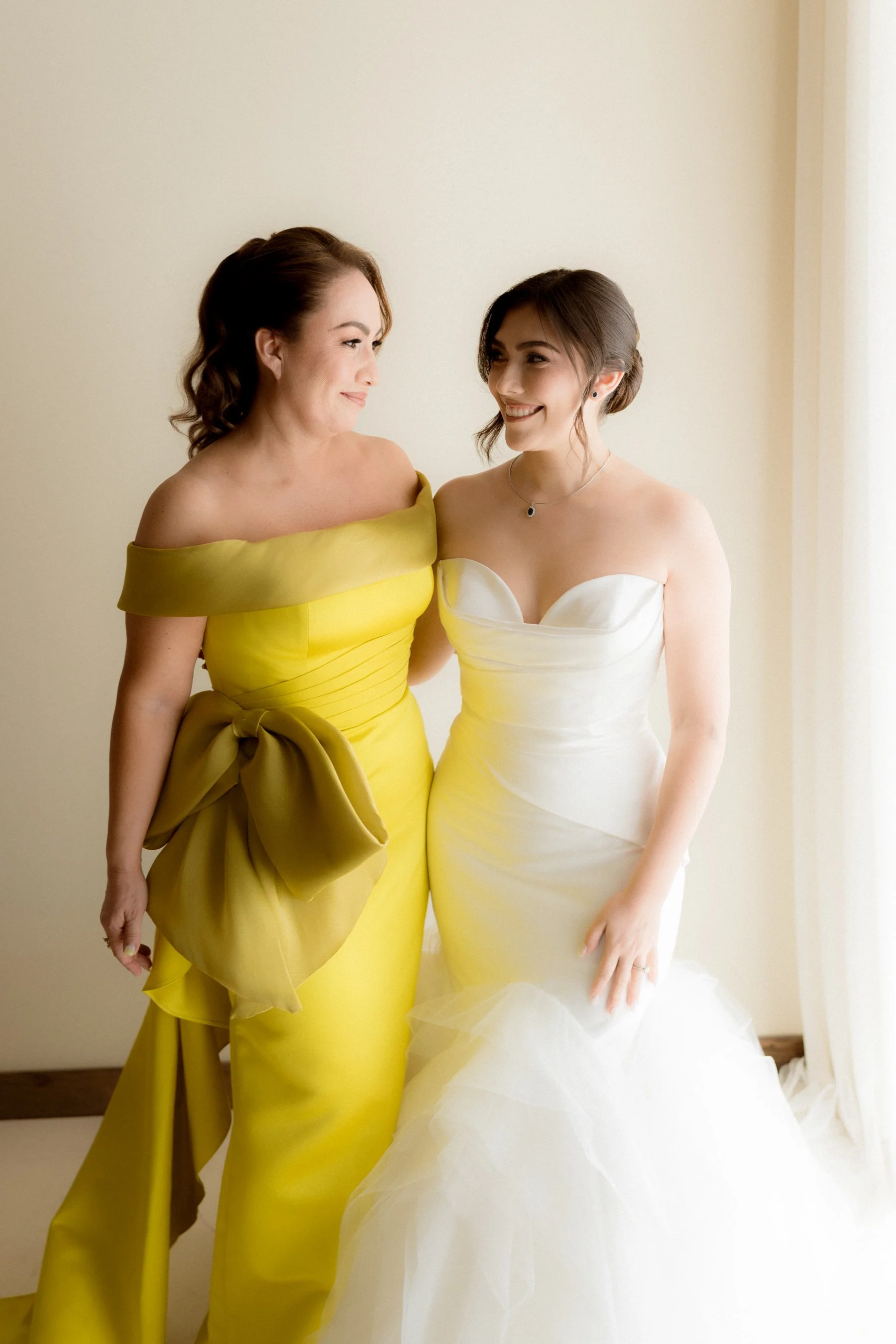 Dos mujeres en ropa de novia y vestido de gala, sonriendo y mirándose en una habitación con luz natural.