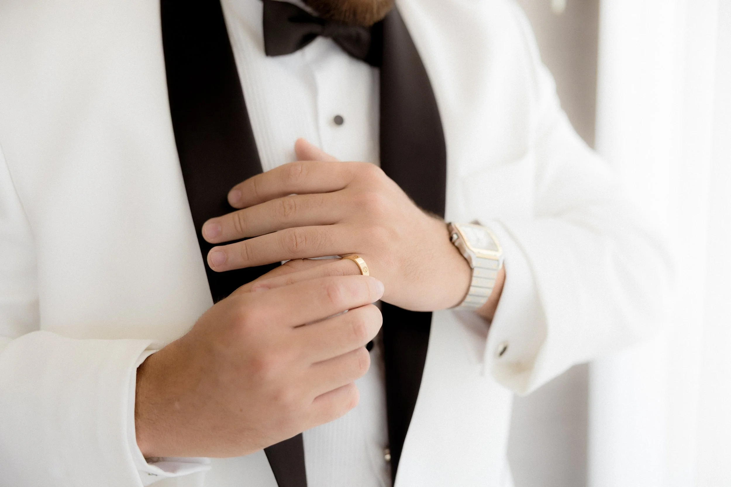 Hombre vestido con esmoquin blanco, usando una chamarra blanca y una pajarita negra, colocando un anillo en su dedo, con un reloj plateado en la muñeca.