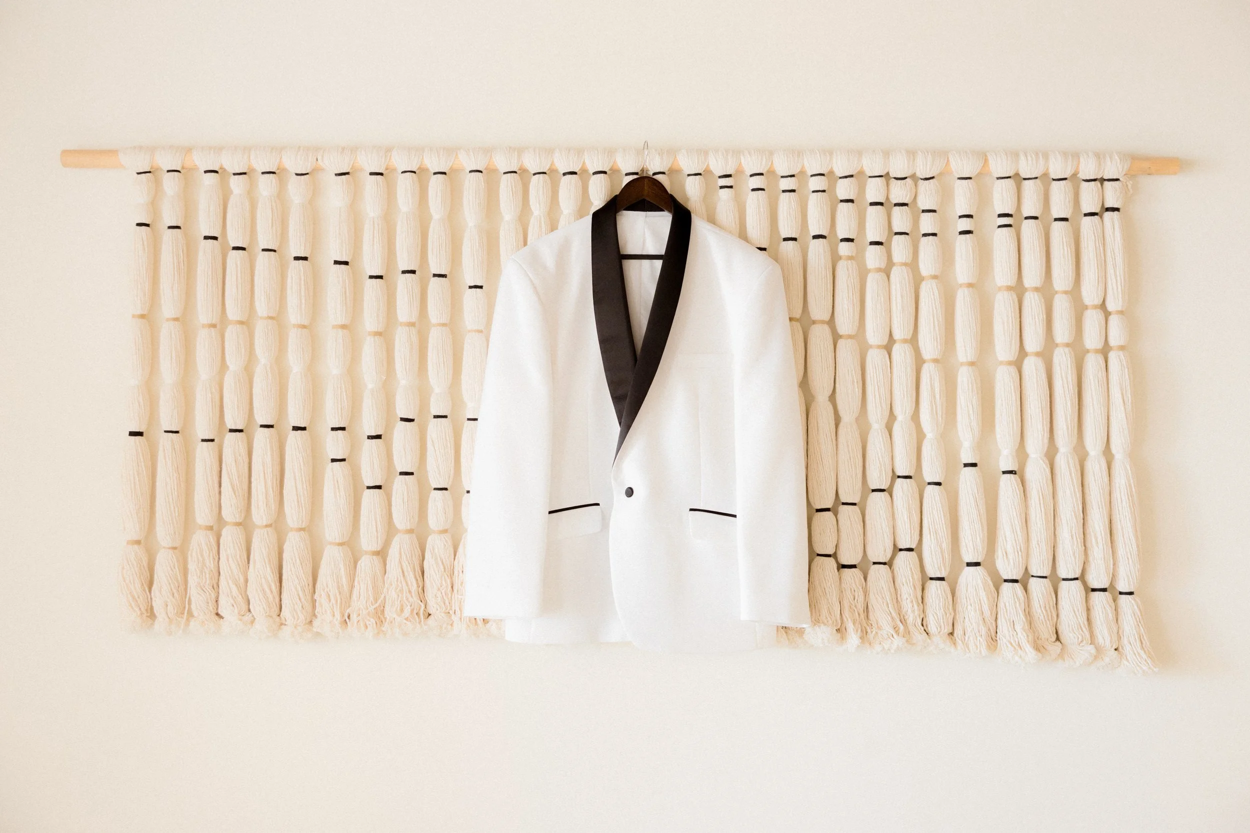 Chaqueta blanca con detalles en negro colgada en un gancho sobre una pared decorativa de cordones blancos con nudos negros.