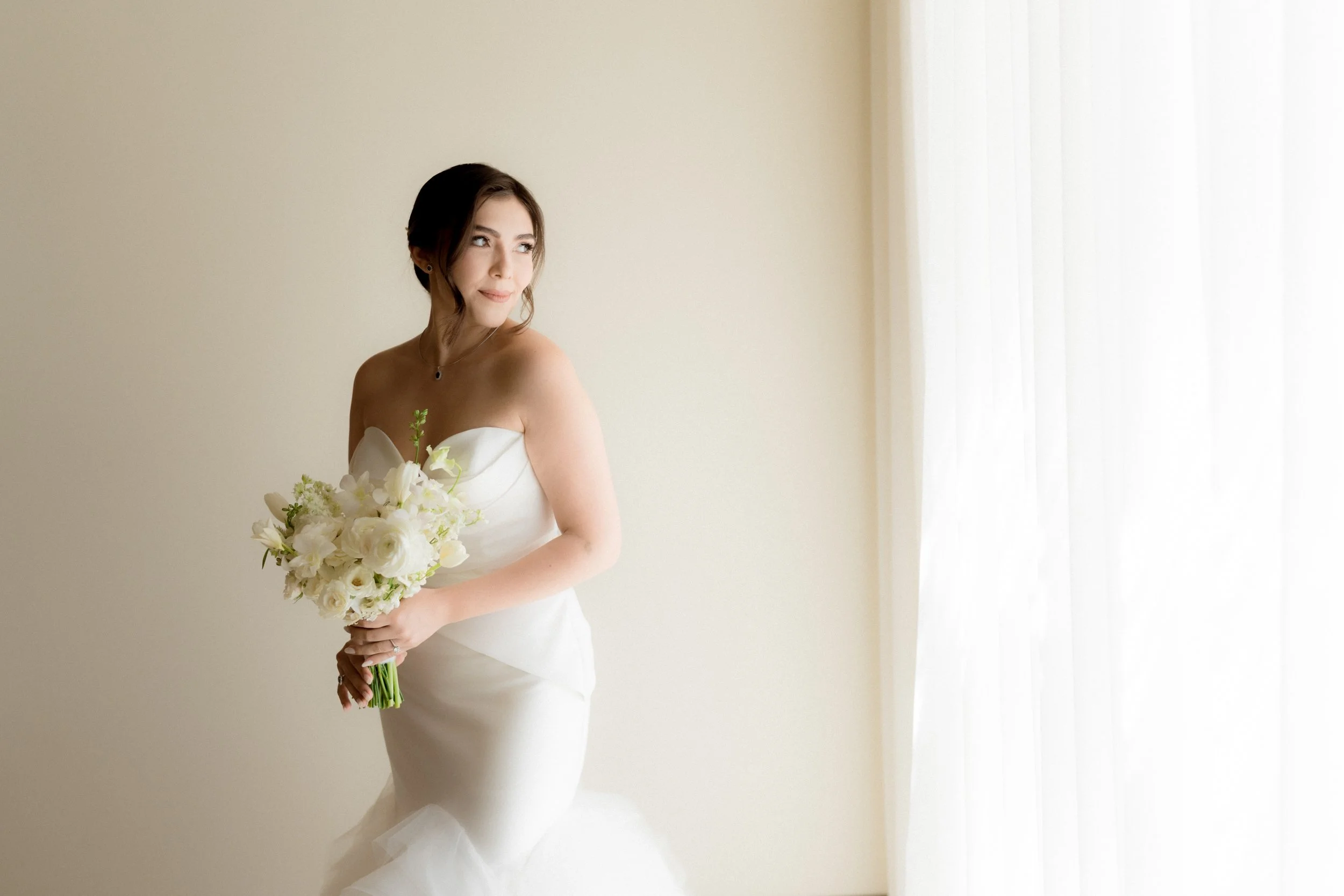 Mujer en vestido de novia sosteniendo un ramo de flores blancas, de perfil, frente a una pared clara y cerca de una ventana con cortinas blancas.