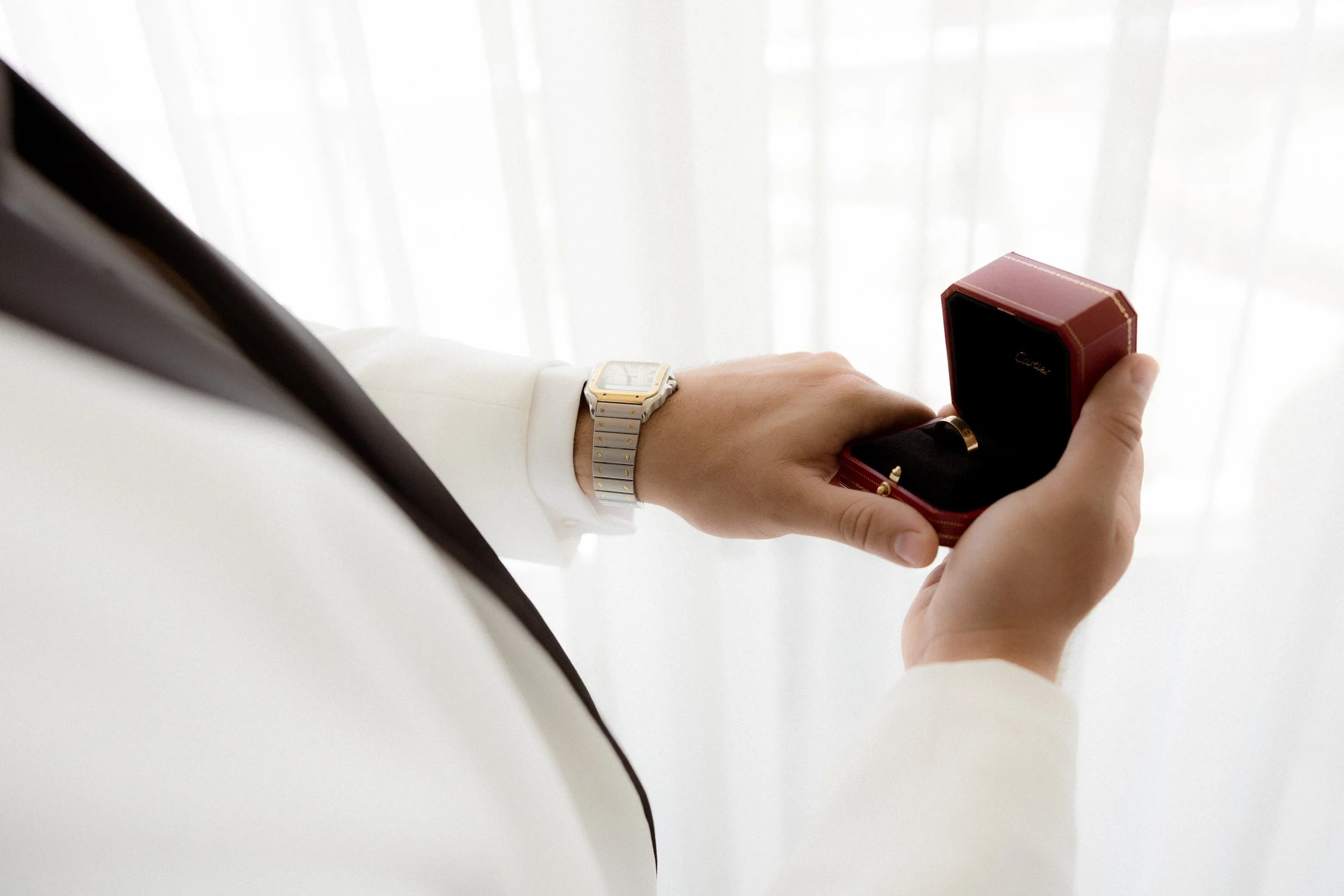 Una persona con traje blanco sosteniedo una caja de reloj con fondo de cortinas blancas.