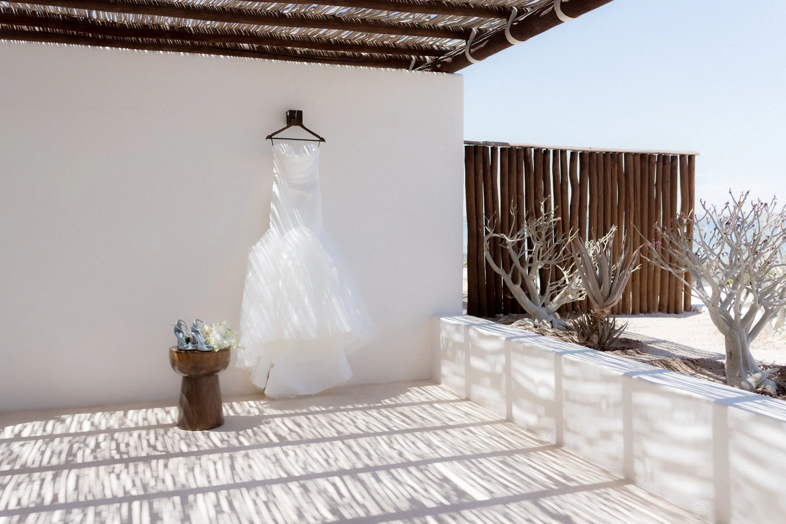 Vestido de boda blanco colgado en una pared blanca, con zapatos de novia y flores en un pequeño banco de madera en un ambiente abierto y soleado con plantas y un muro de madera.